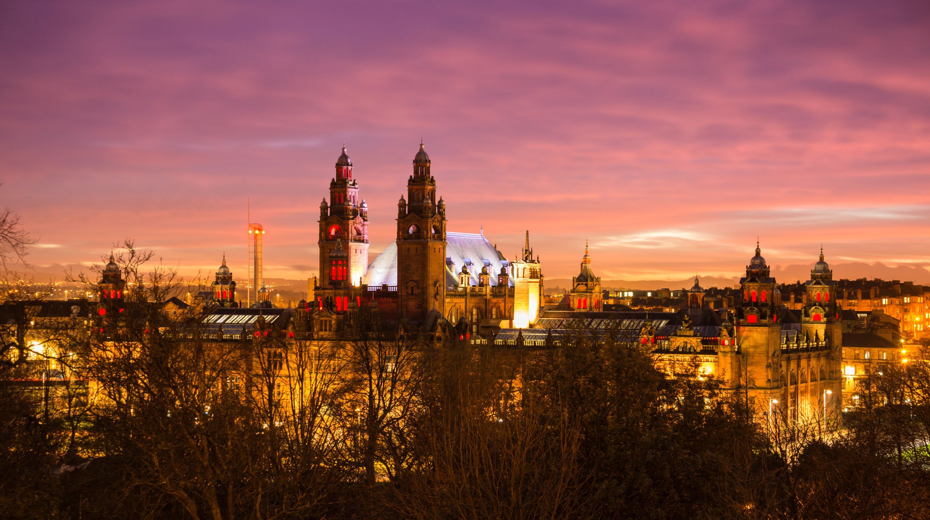 The Glasgow University building is illuminated at dusk with a purple and orange sky above and silhouetted trees in the foreground.