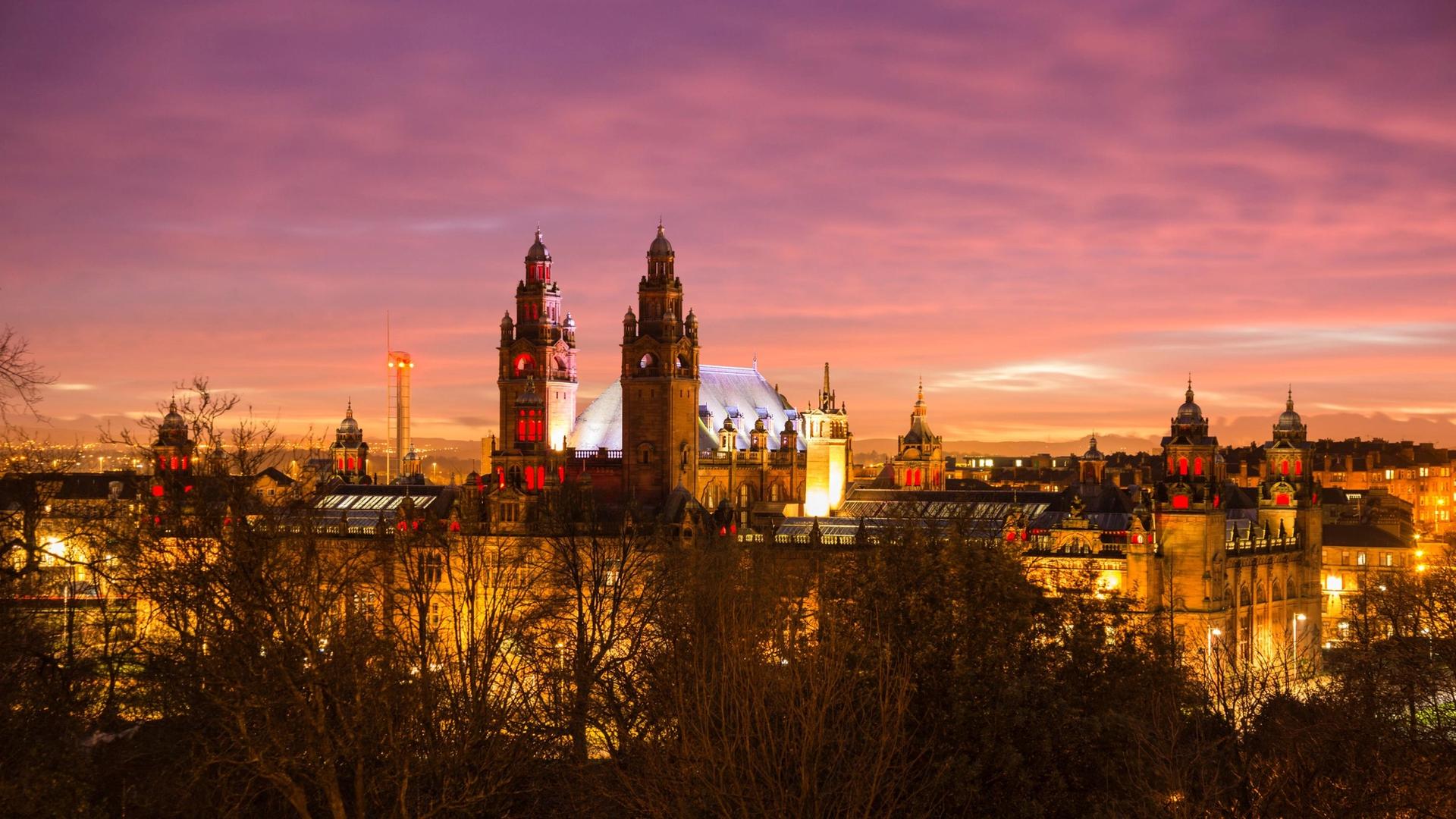The Glasgow University building is illuminated at dusk with a purple and orange sky above and silhouetted trees in the foreground.