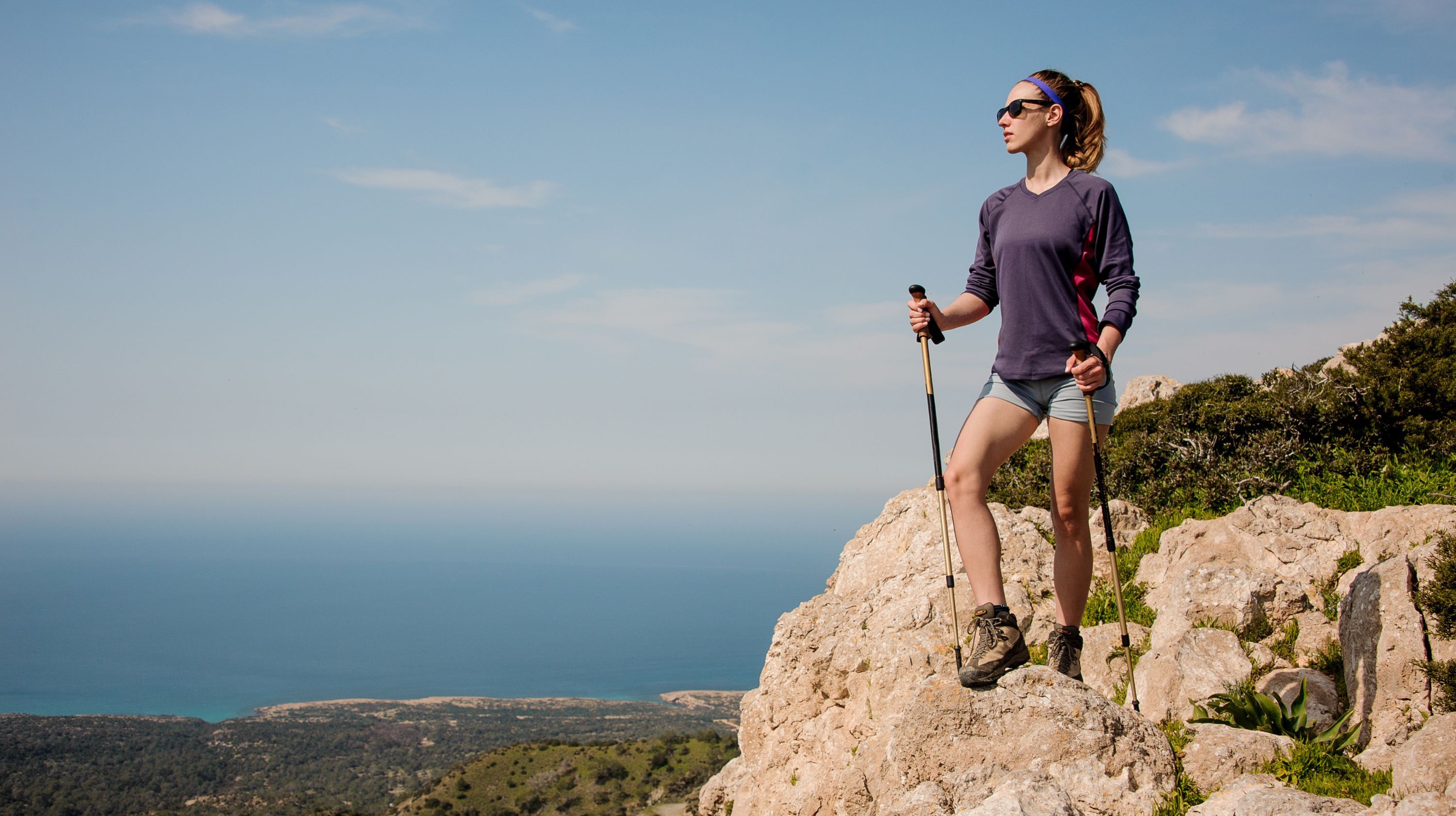 A person stands on a rocky cliff overlooking the ocean, holding trekking poles. They are wearing sunglasses and a purple long-sleeved shirt with shorts.