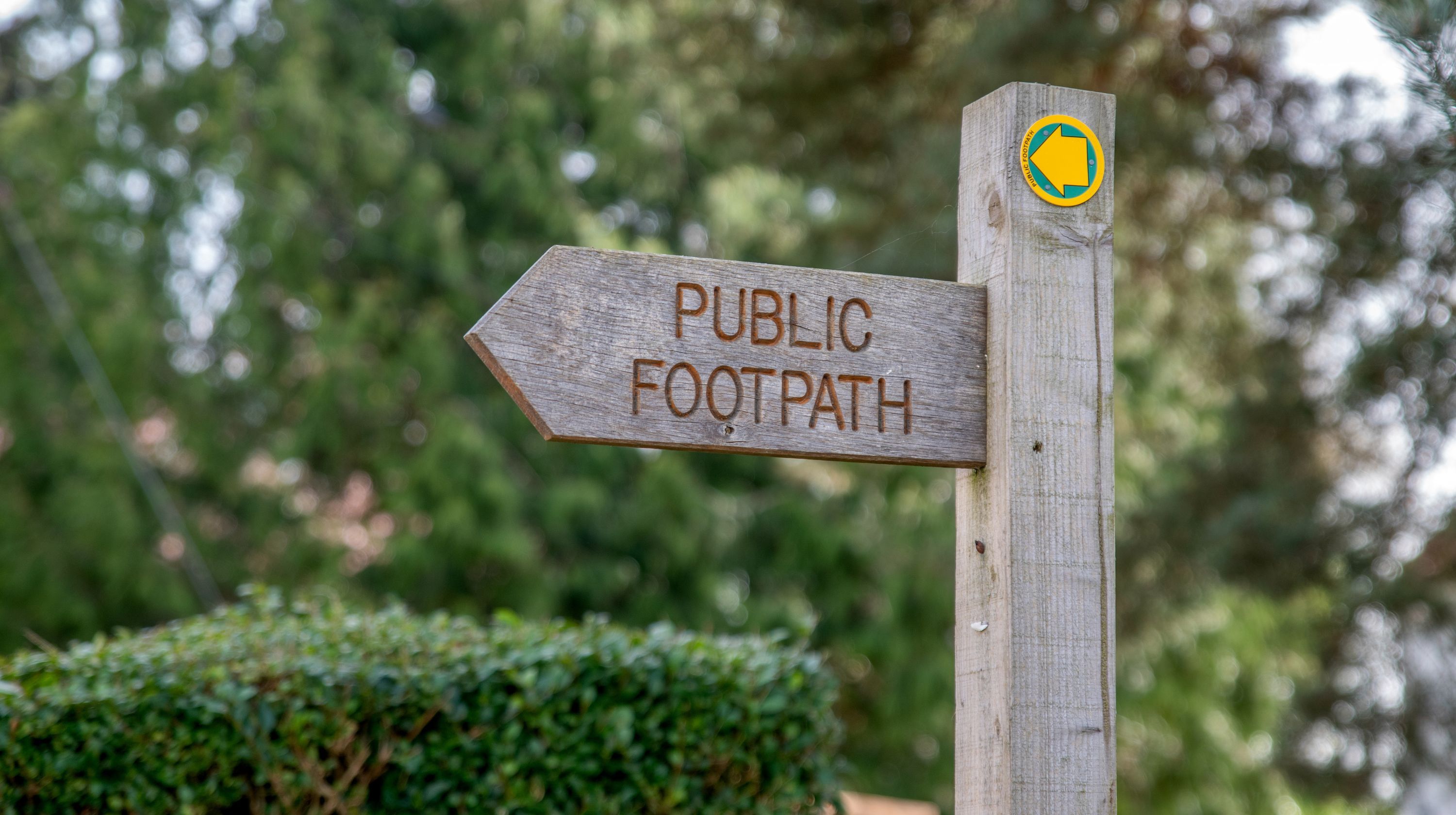 A wooden signpost indicates a public footpath with an arrow pointing left. A yellow circular marker with a left-pointing arrow is attached to the post.