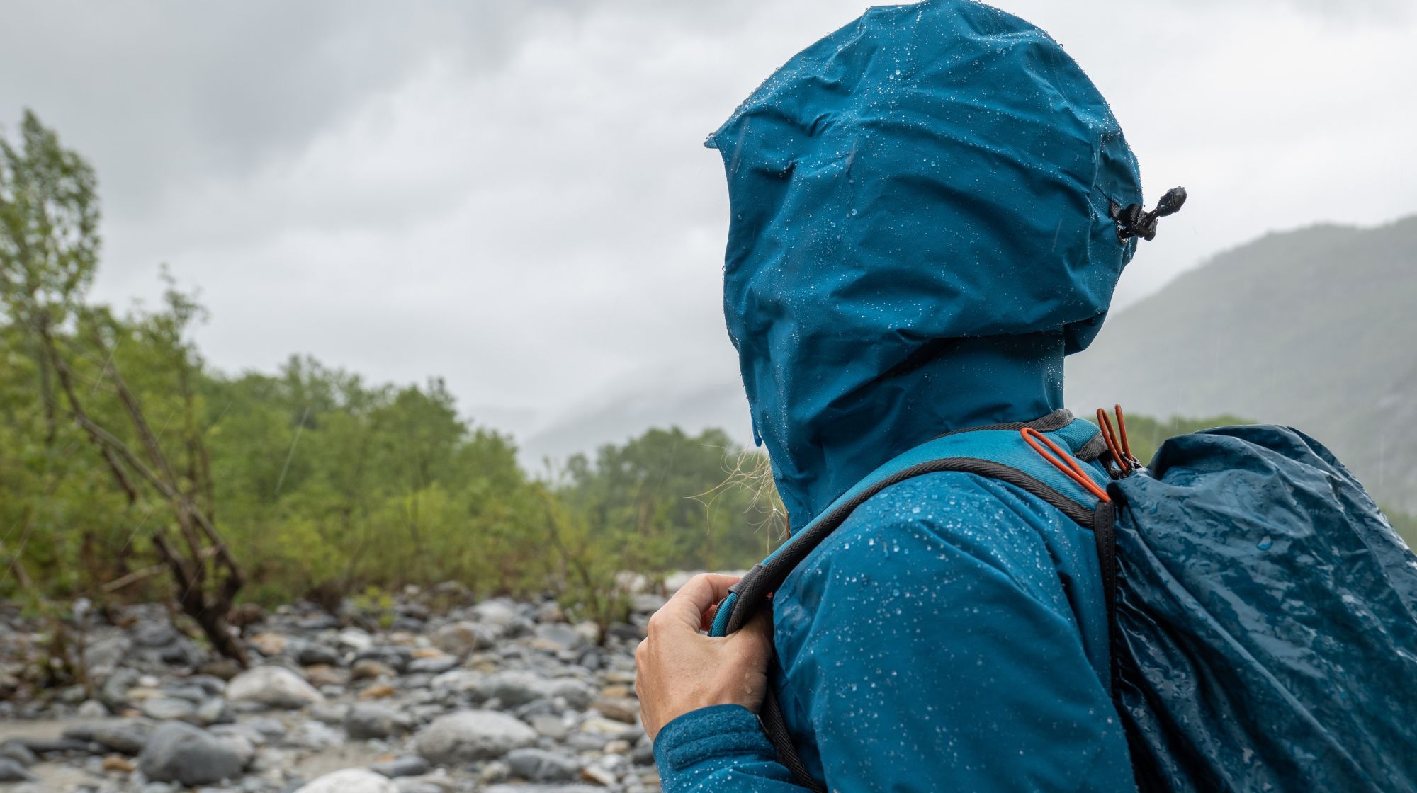 A person in a hooded blue jacket and backpack stands in a rocky landscape on a cloudy day