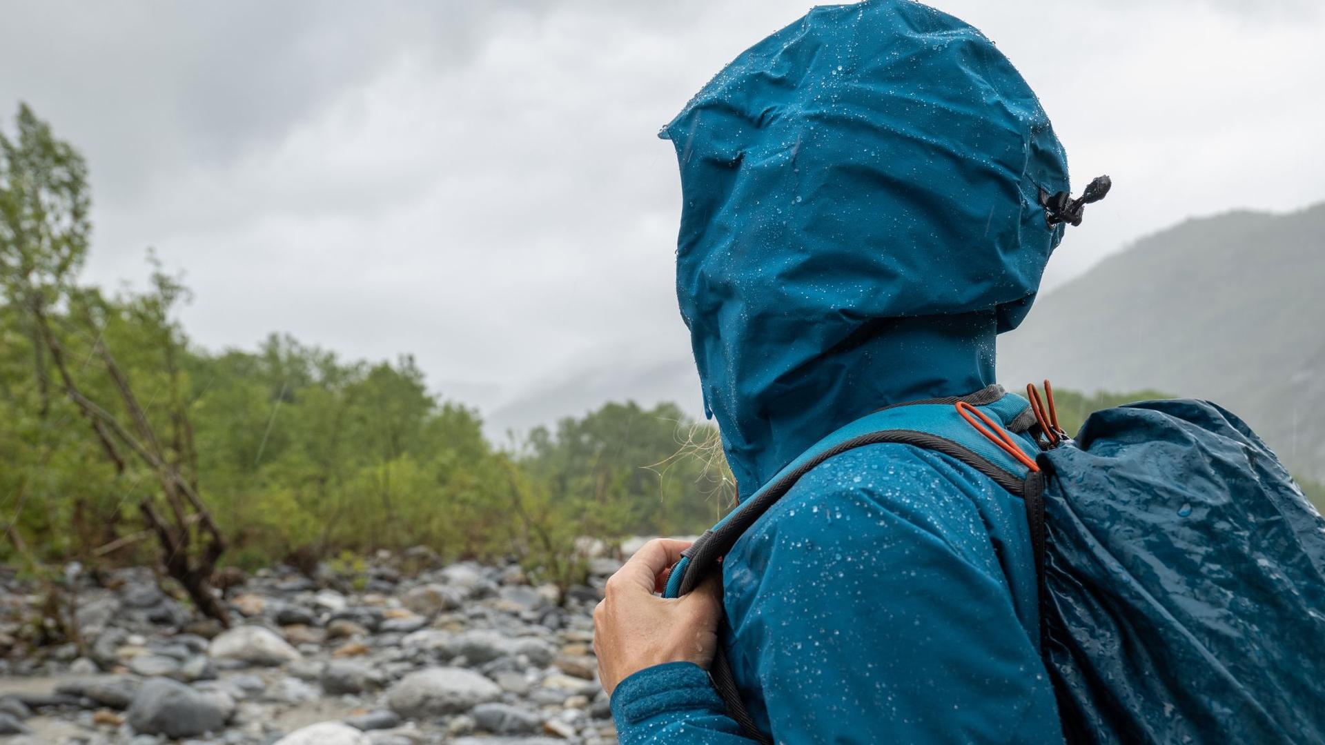 A person in a hooded blue jacket and backpack stands in a rocky landscape on a cloudy day