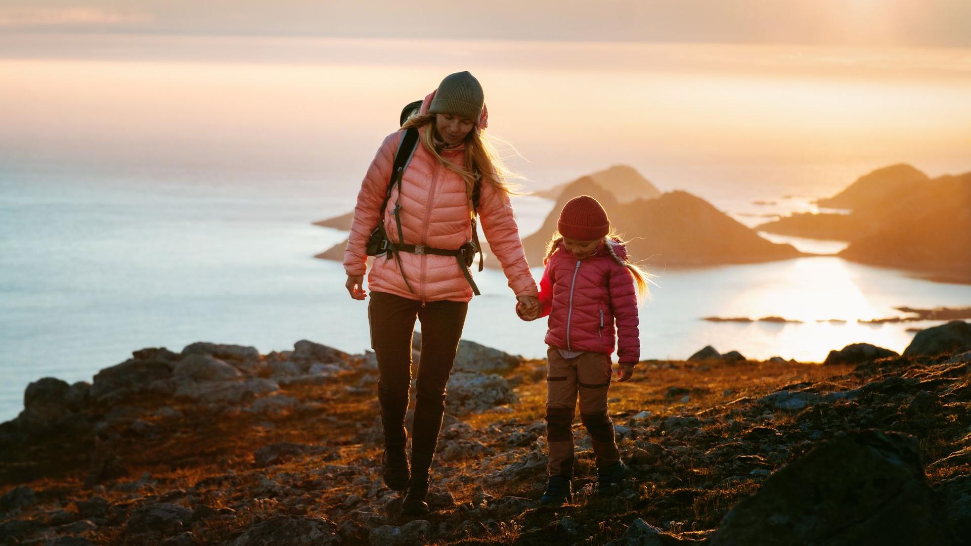 Two hikers, one adult and one child, hold hands while walking on a rocky trail overlooking a bay during sunset.