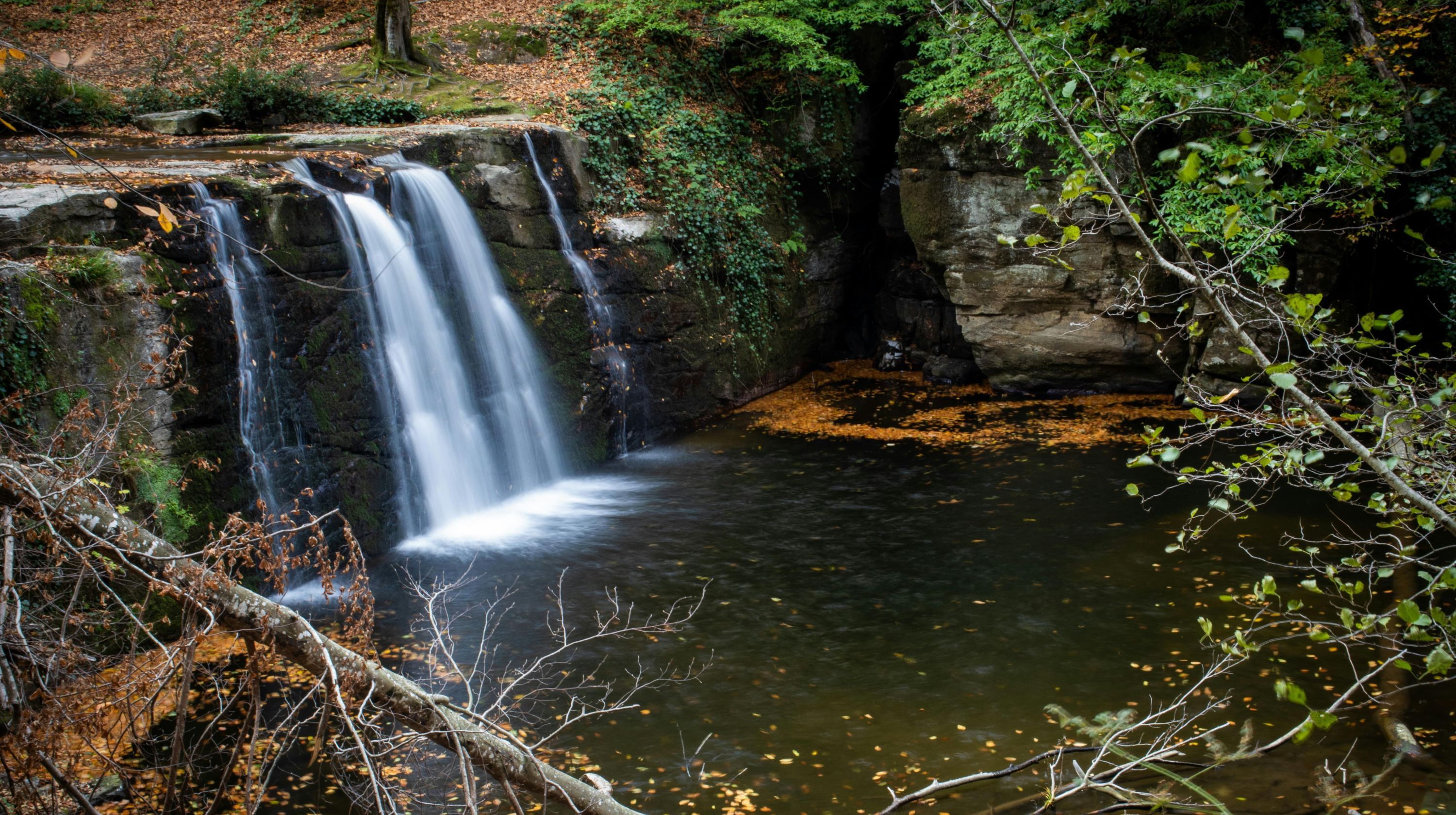 A serene waterfall cascades down mossy rocks into a dark pool, surrounded by autumn foliage and lush green trees.