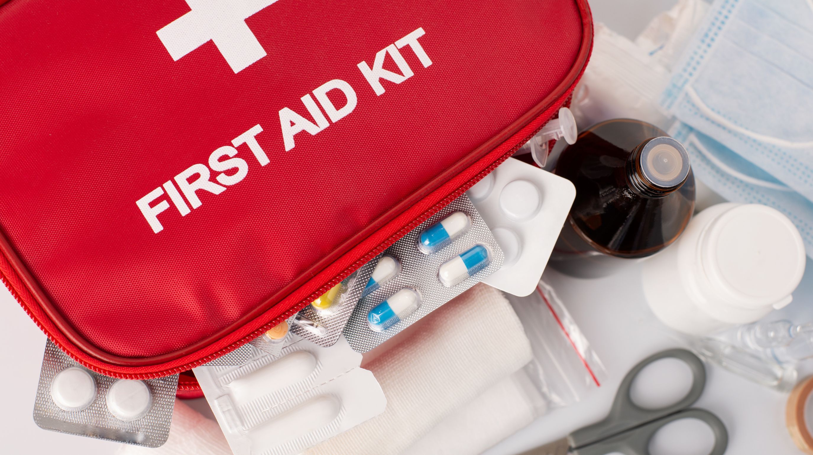 A red first aid kit bag is open, revealing various medical supplies including blister packs of pills, a small bottle of liquid, a white jar, bandages, and scissors.