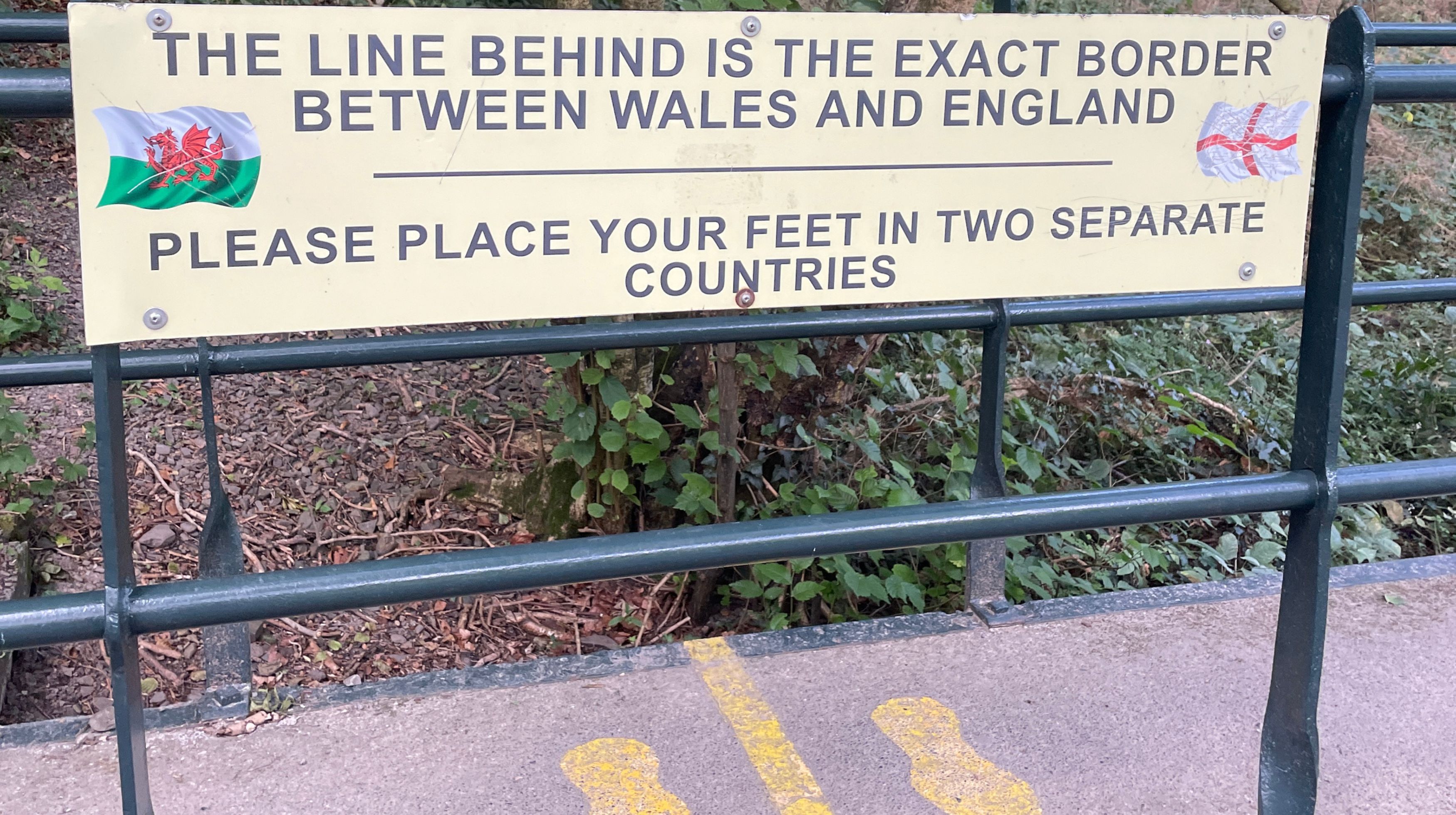 A sign reads: "The line behind is the exact border between Wales and England. Please place your feet in two separate countries." The flags of Wales and England are shown on the sign.