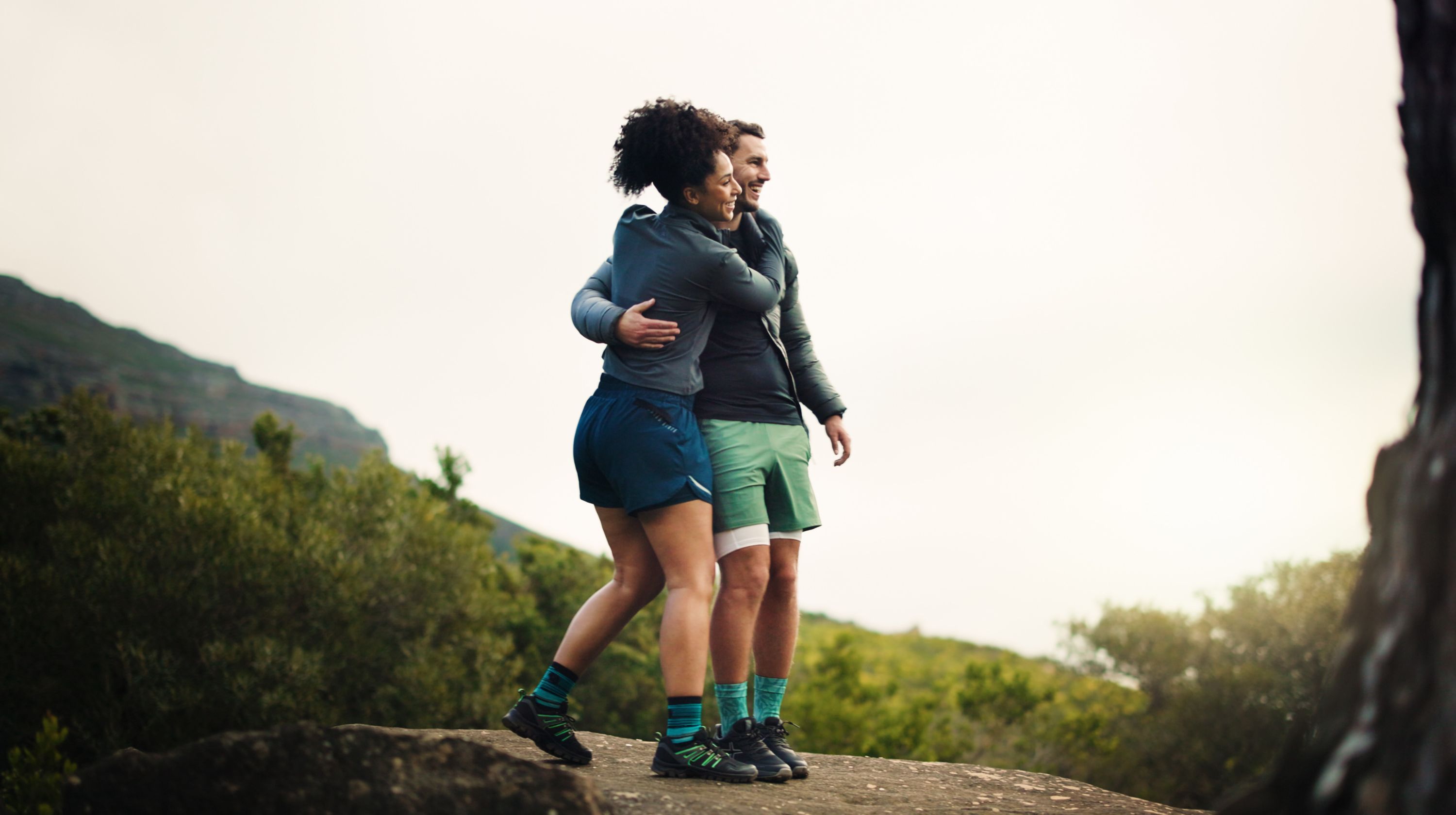 Two people in athletic wear embracing on a rocky outcrop with lush green foliage and distant mountains under a bright, overcast sky.