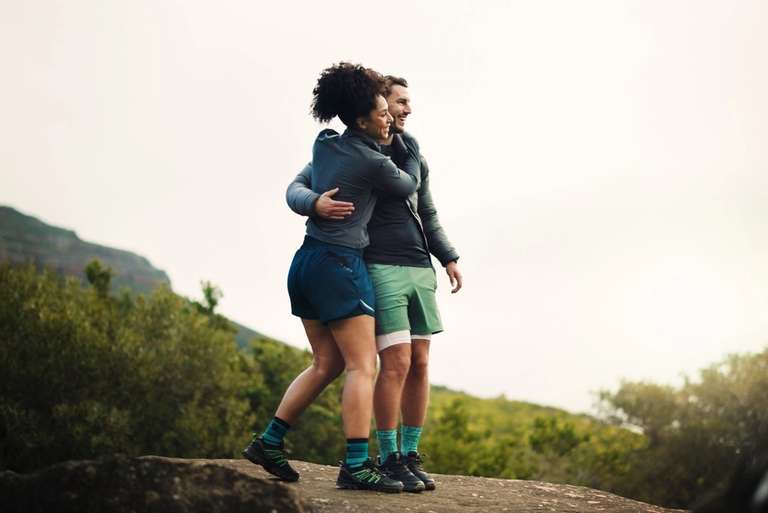 Two people in athletic wear embracing on a rocky outcrop with lush green foliage and distant mountains under a bright, overcast sky.