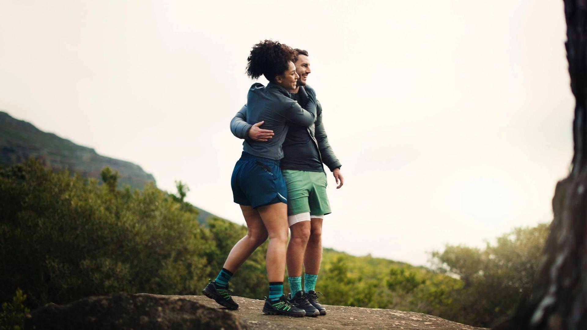 Two people in athletic wear embracing on a rocky outcrop with lush green foliage and distant mountains under a bright, overcast sky.