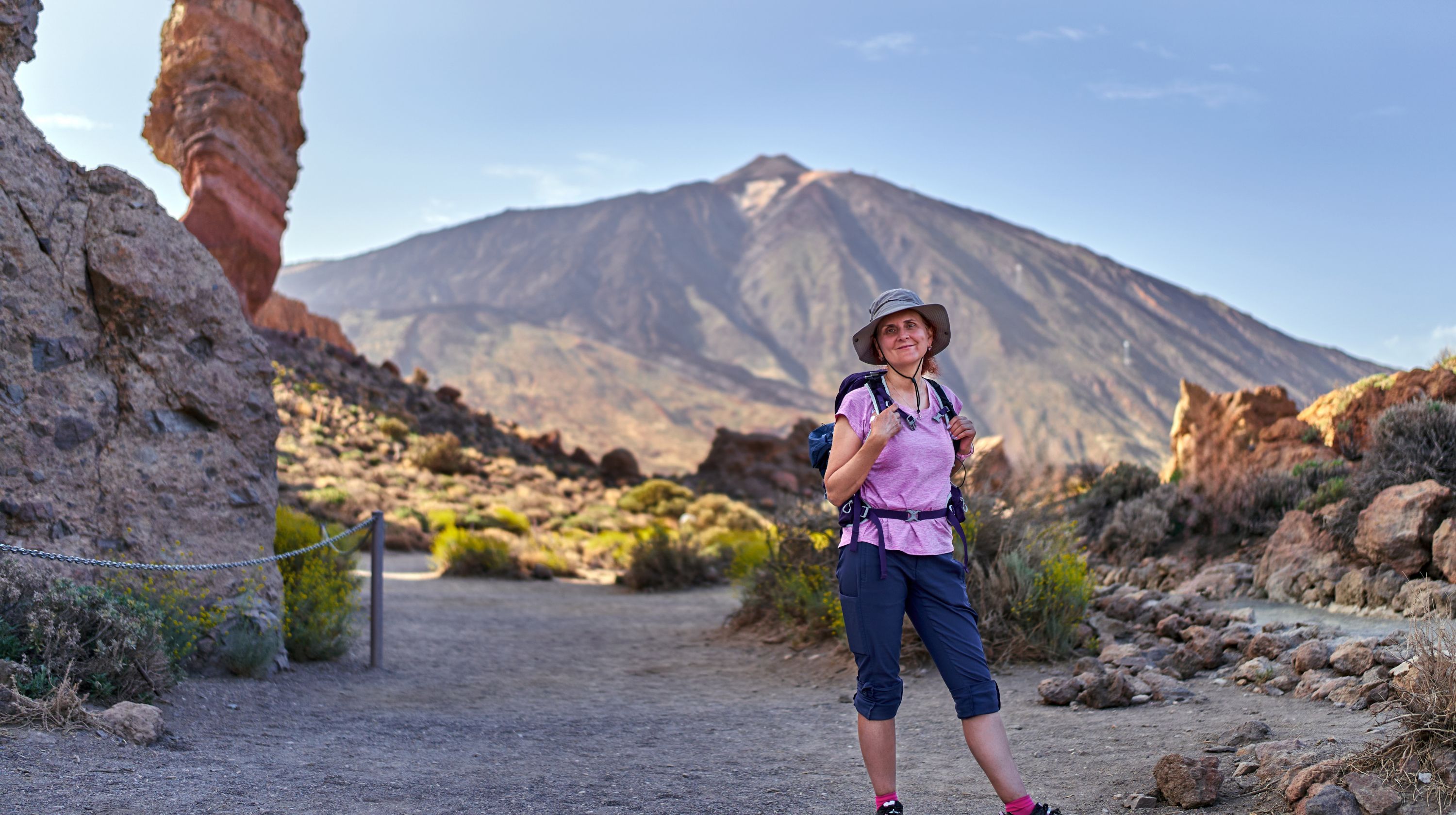 A hiker stands on a path in a rocky, mountainous landscape with a volcano in the background.
