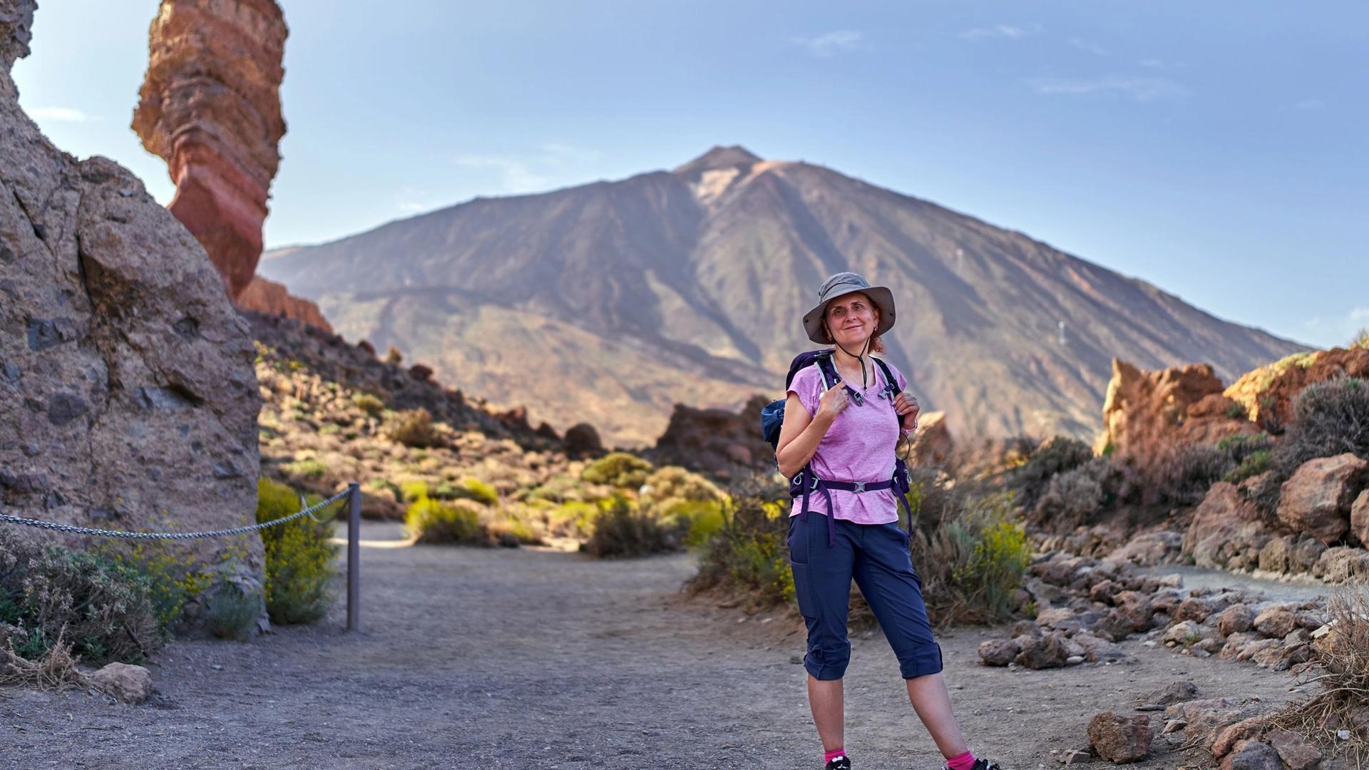 A hiker stands on a path in a rocky, mountainous landscape with a volcano in the background.