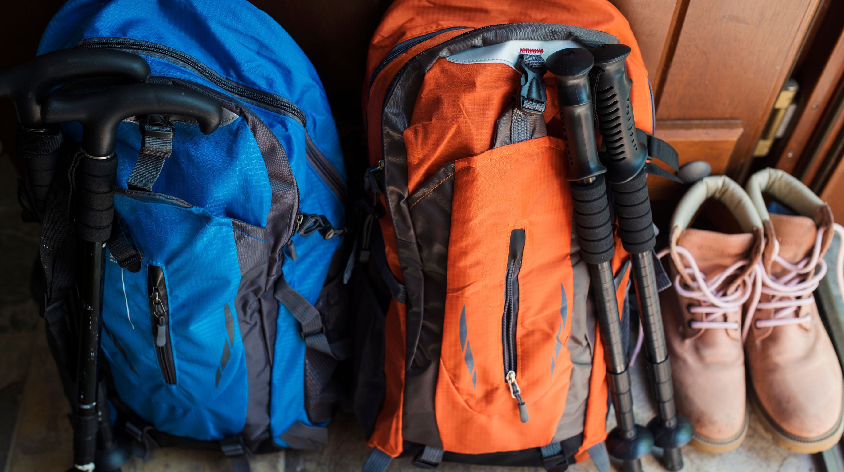 Two backpacks, one blue and one orange, stand next to a pair of tan hiking boots and trekking pole