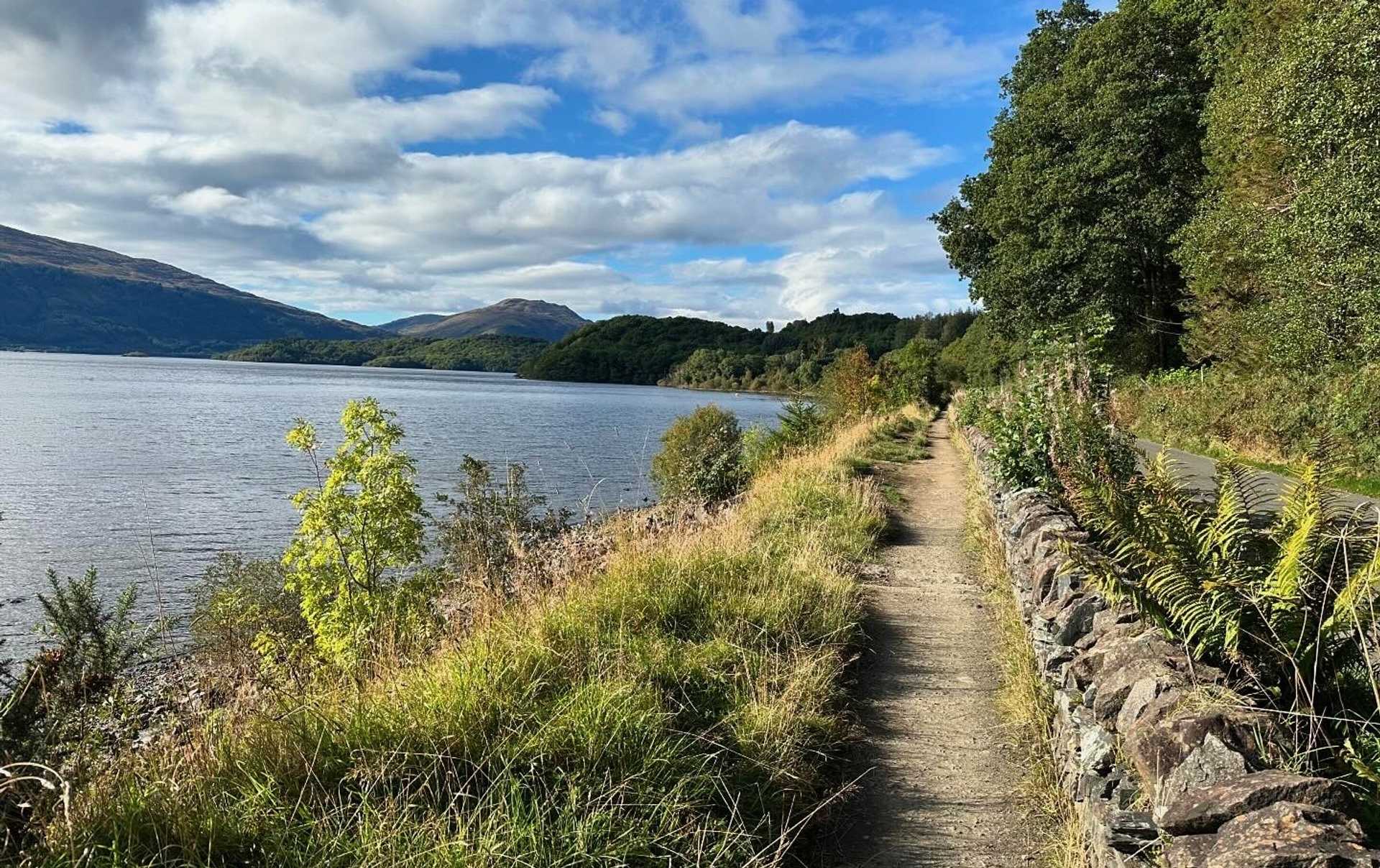 Narrow West Highland Way path following the eastern shore of Loch Lomond, with hills beyond.