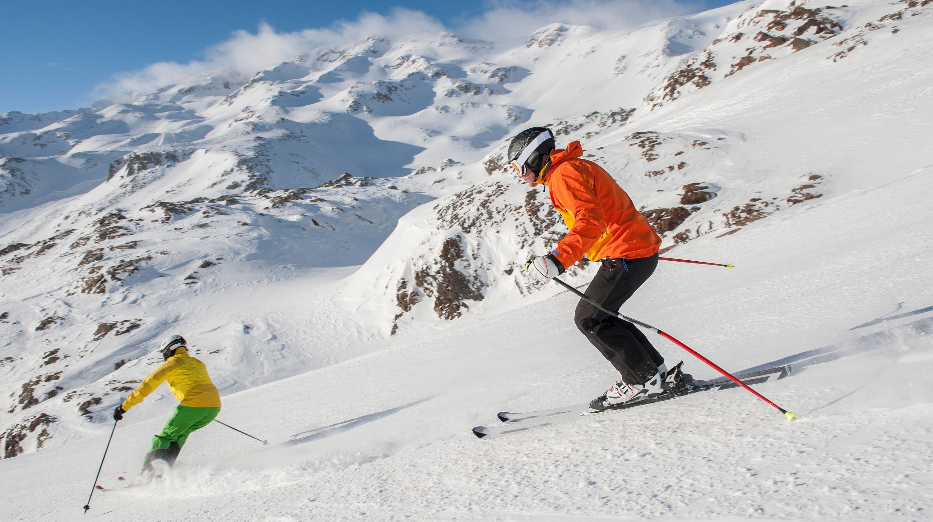 Two skiers skiing down a snow-covered mountain under a bright blue sky with clouds.