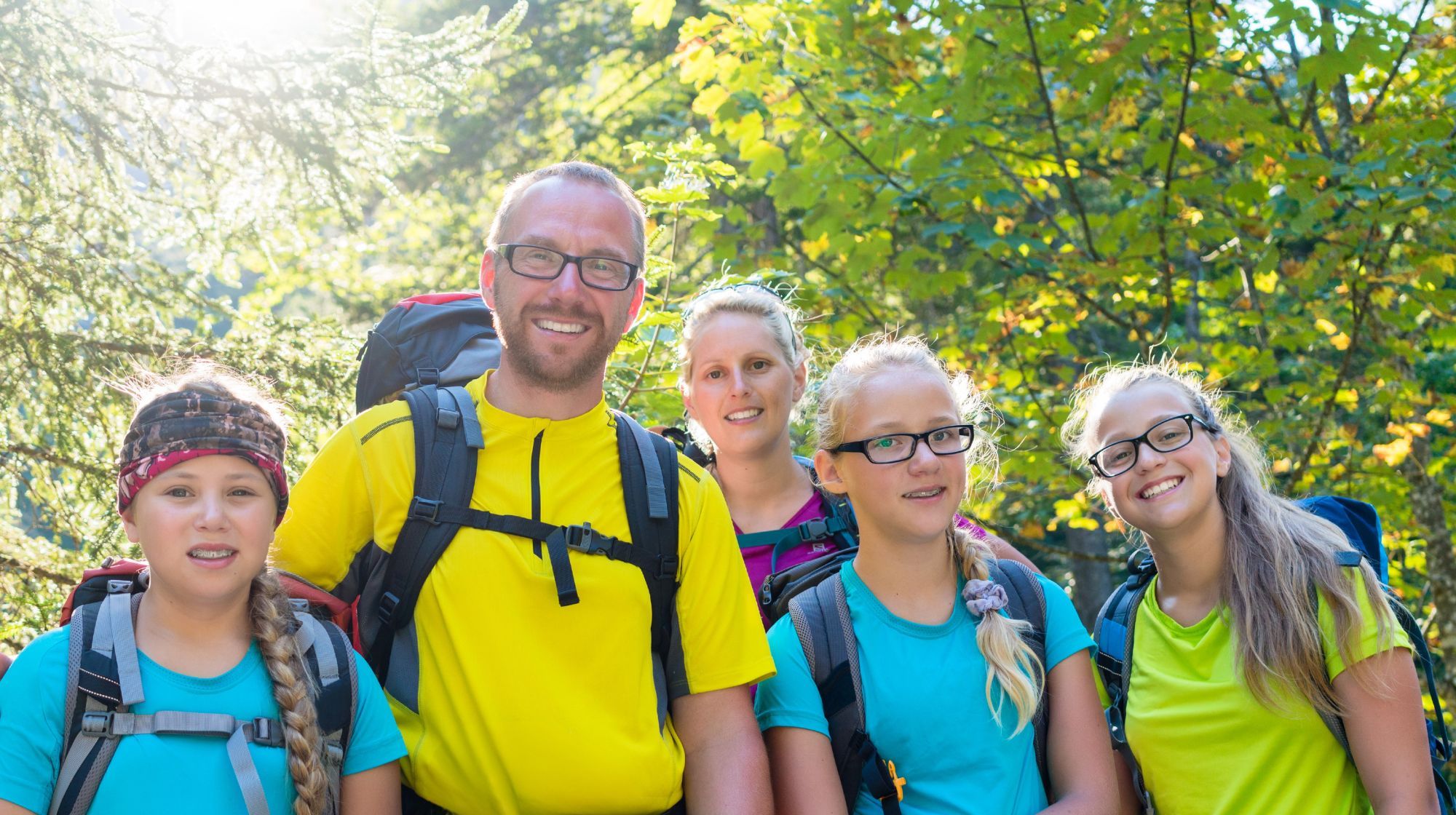 A group of five people, including children, wearing backpacks and smiling in a sun-dappled forest.