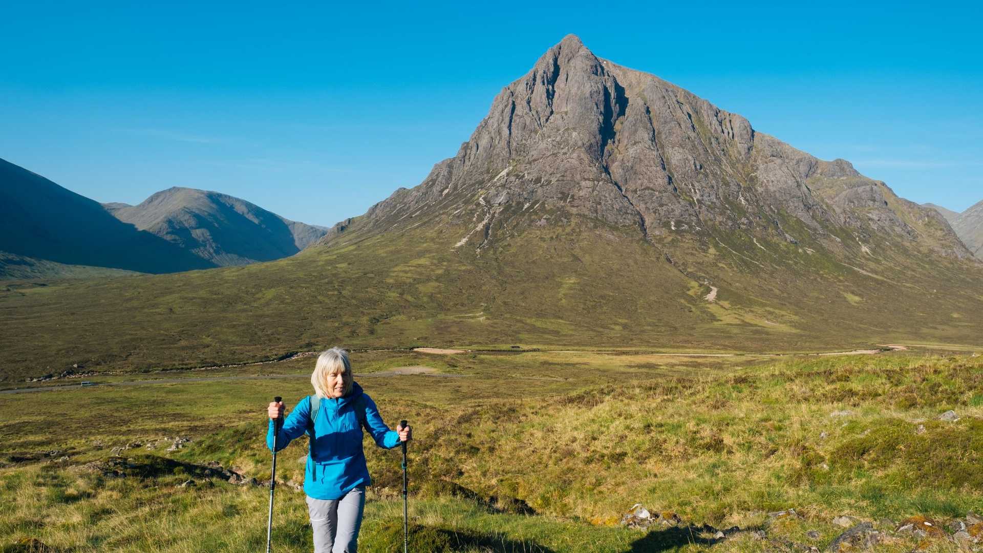 A person hikes on a grassy mountain trail with a large, rugged mountain peak in the background under a clear blue sky.