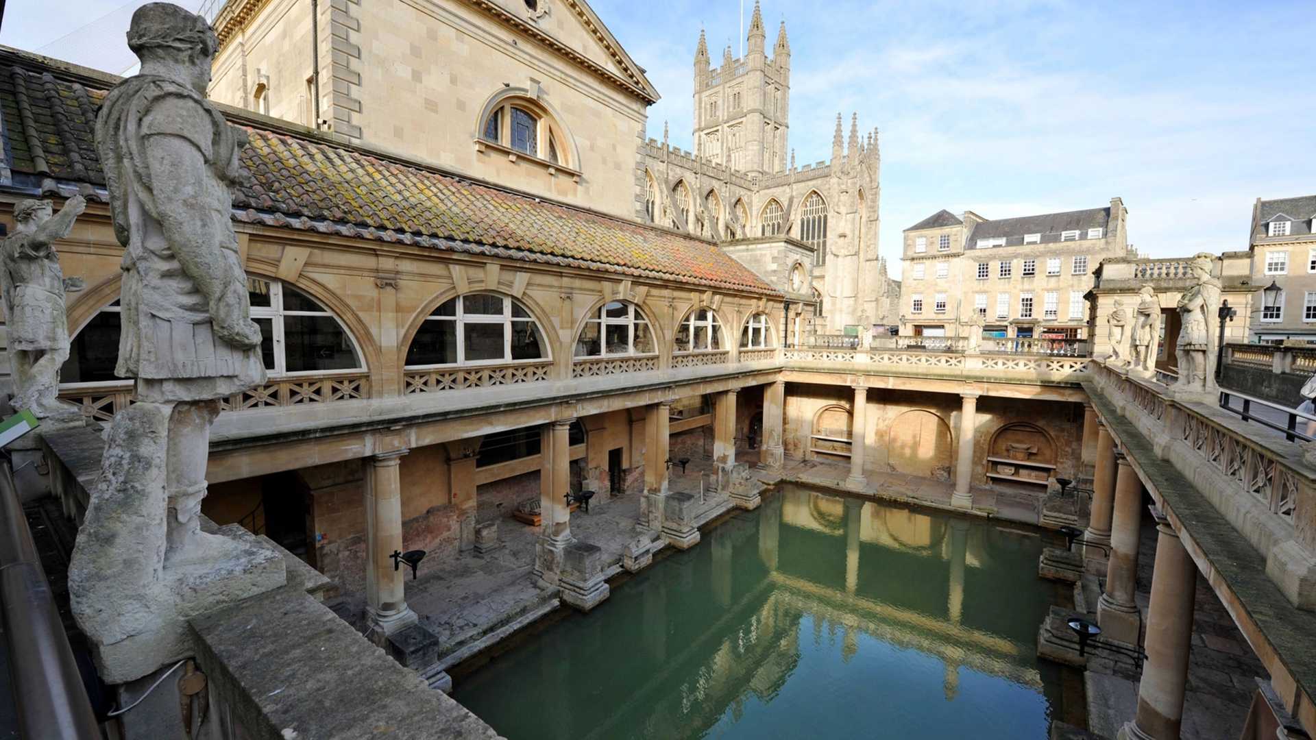The Roman Baths in Bath, England, feature a large, green pool surrounded by ancient stone architecture and statues, with Bath Abbey visible in the background.