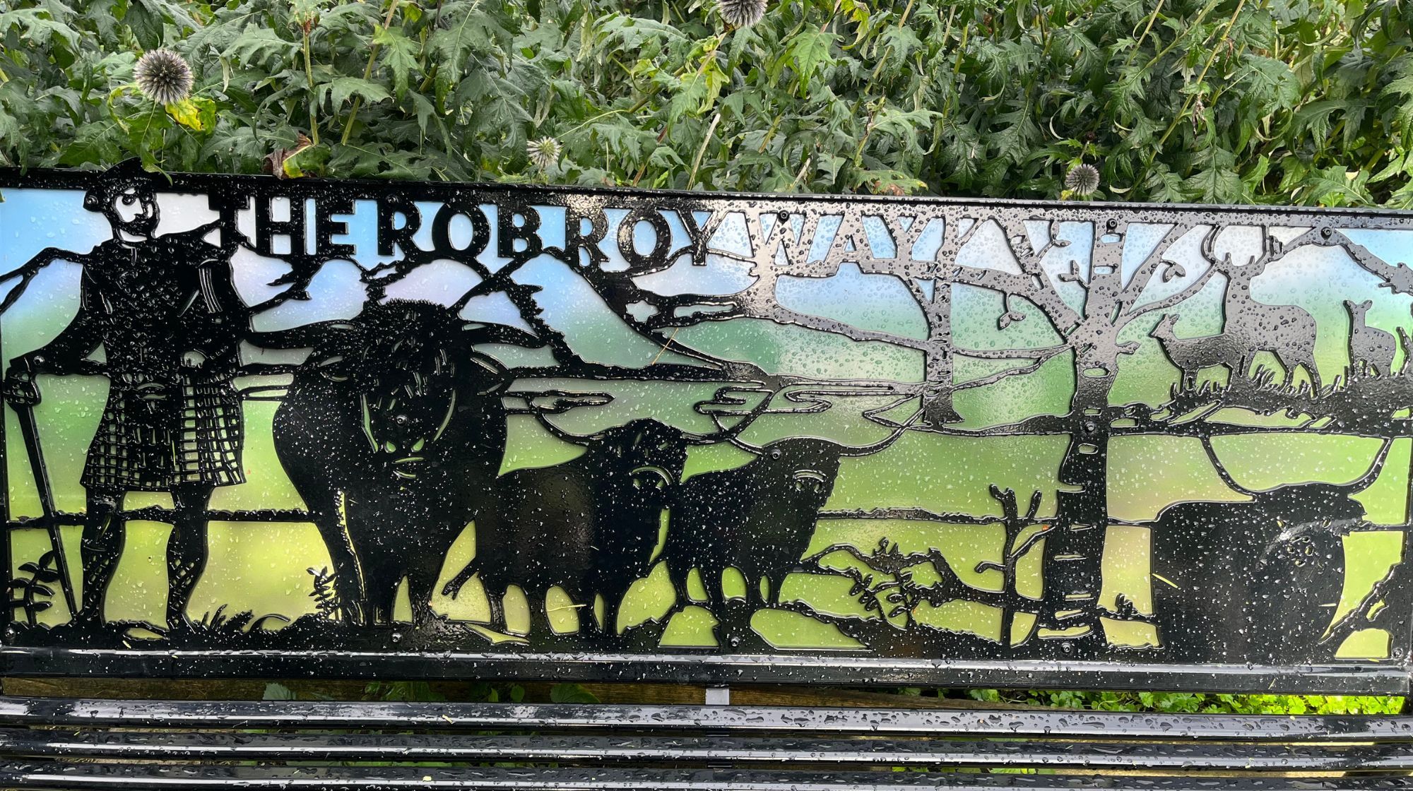 A metal bench with "THE ROB ROY WAY" inscribed, featuring silhouettes of a kilted man, highland cows, deer, and trees against a scenic backdrop.