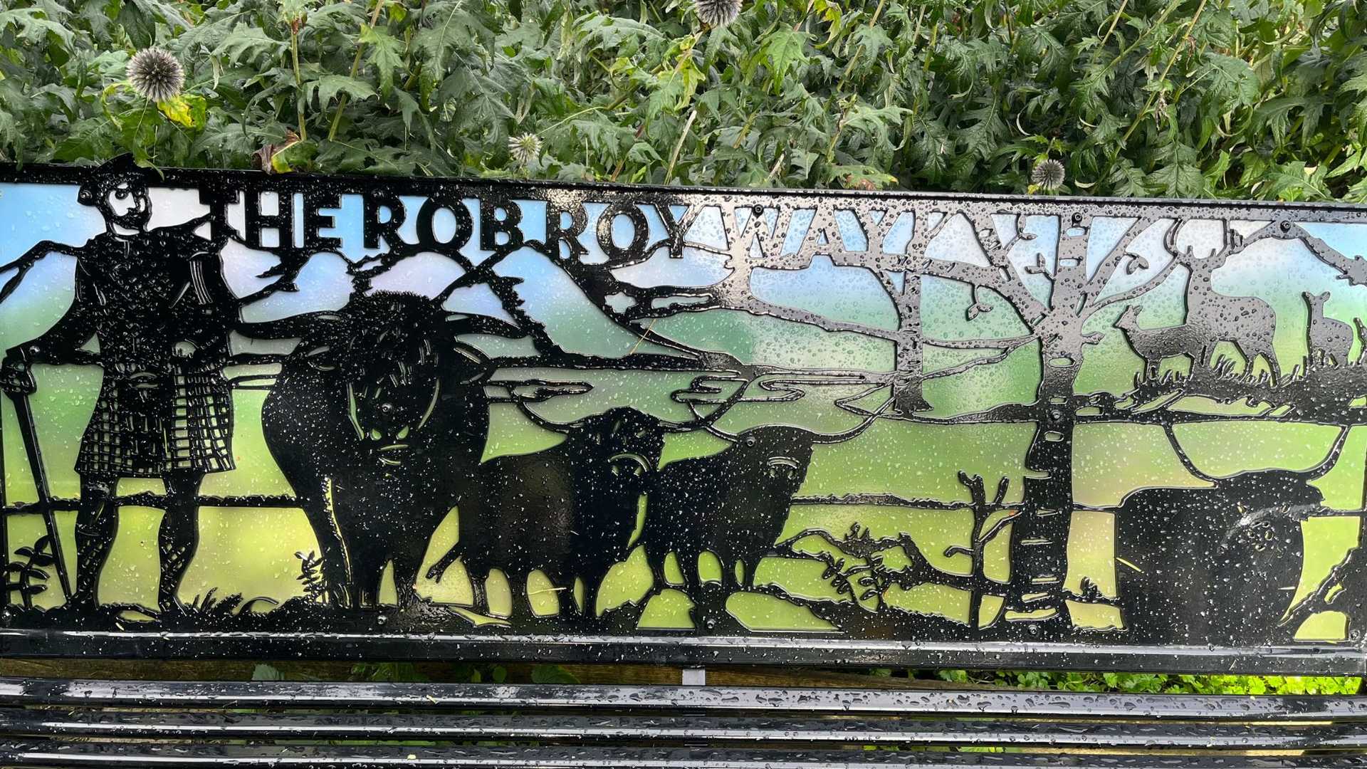 A metal bench with "THE ROB ROY WAY" inscribed, featuring silhouettes of a kilted man, highland cows, deer, and trees against a scenic backdrop.