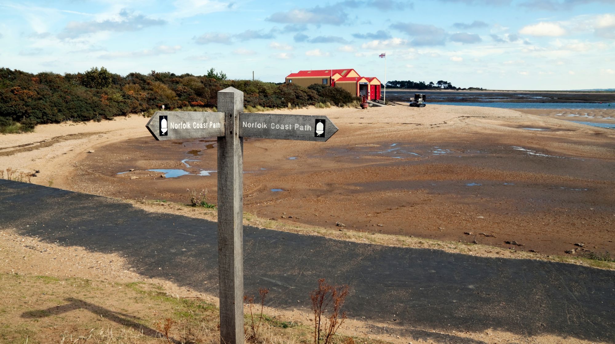 A wooden signpost with arrows pointing left and right, indicating the Norfolk Coast Path, stands on a sandy beach with a body of water in the background and a red-roofed building further inland.
