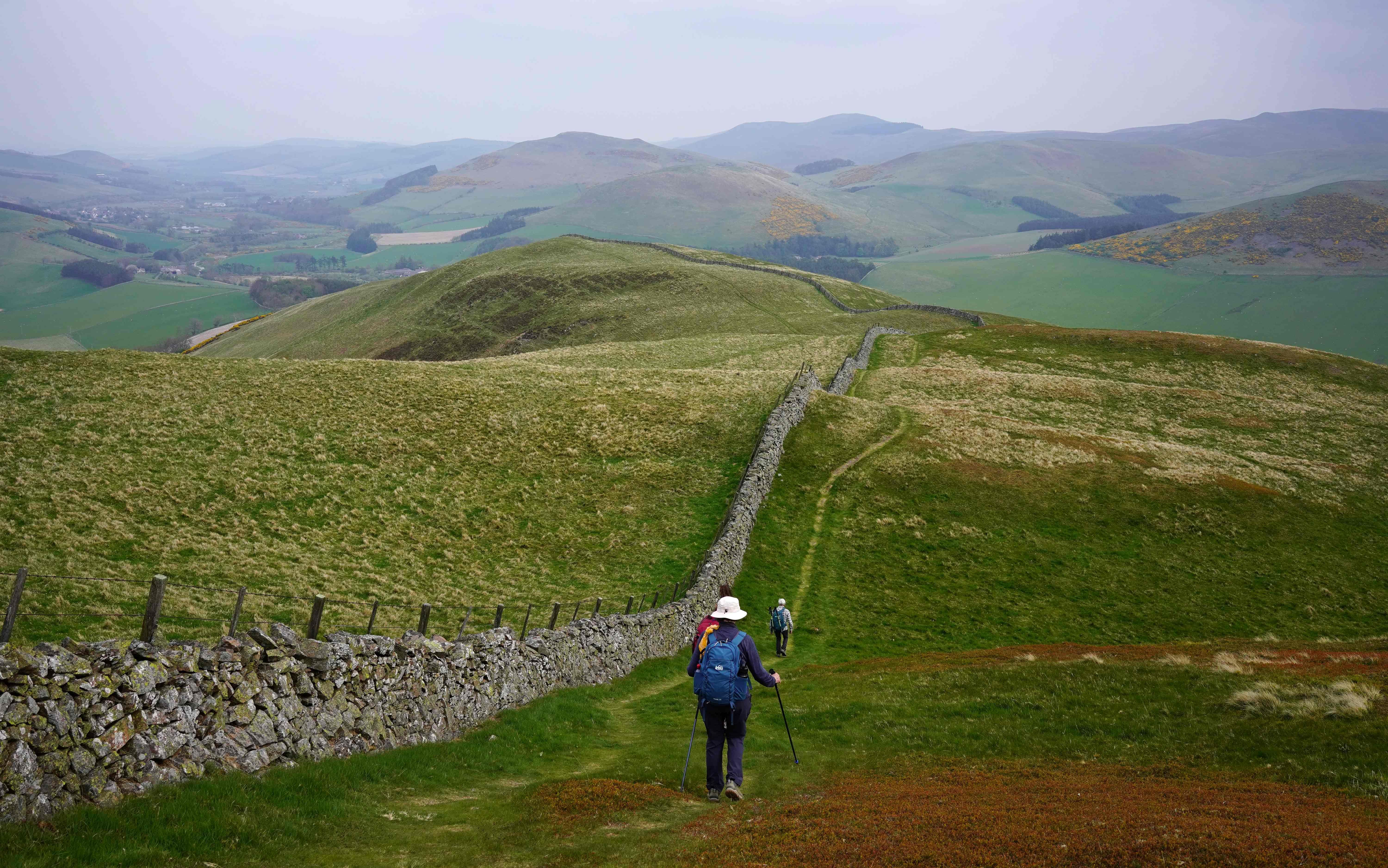 Two hikers walk along a grassy path next to a stone wall, with rolling green hills and a valley in the background.
