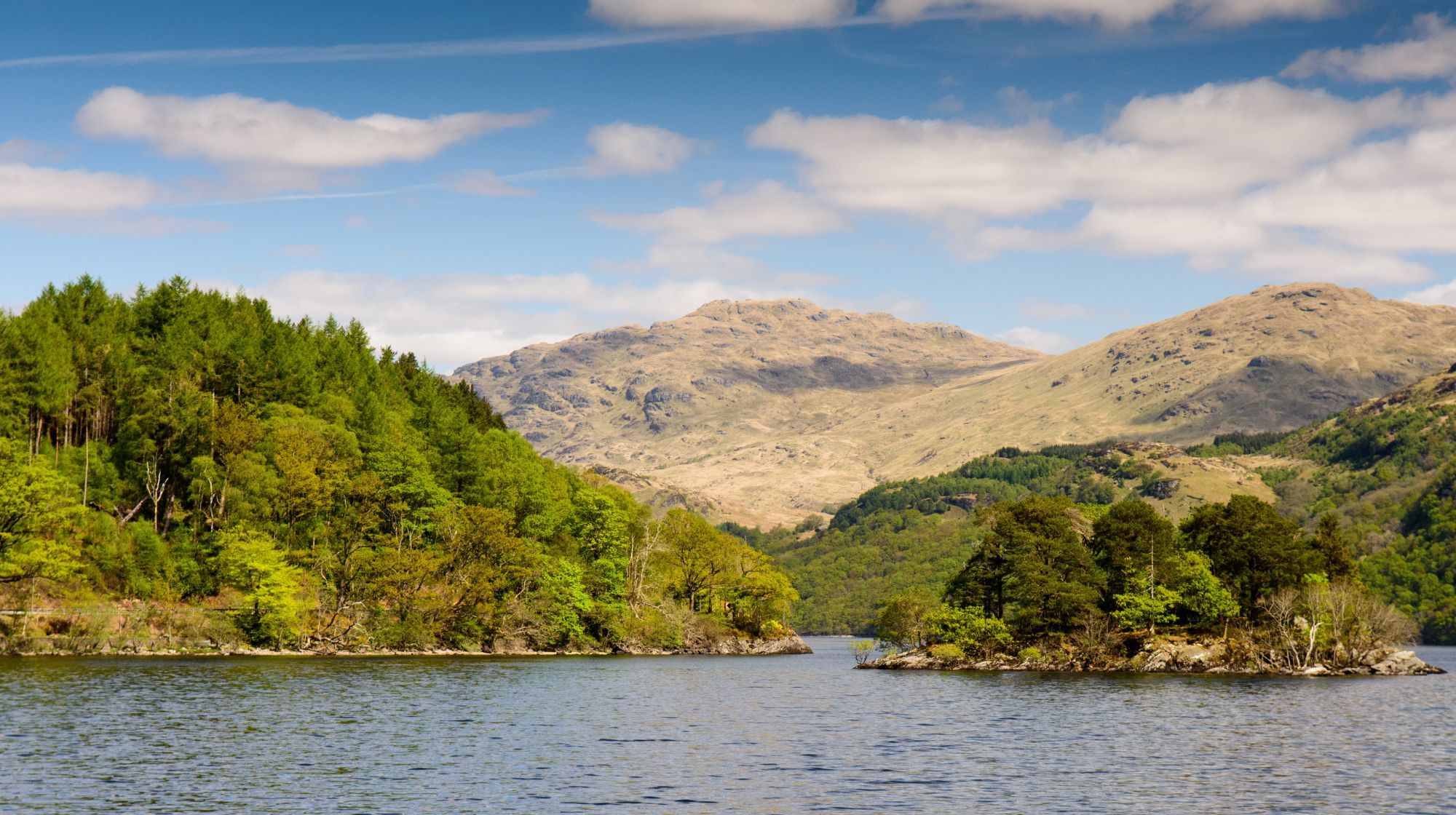 A wide body of water with a small, tree-covered island in the foreground, leading to lush green hills and barren, rocky mountains under a cloudy blue sky.
