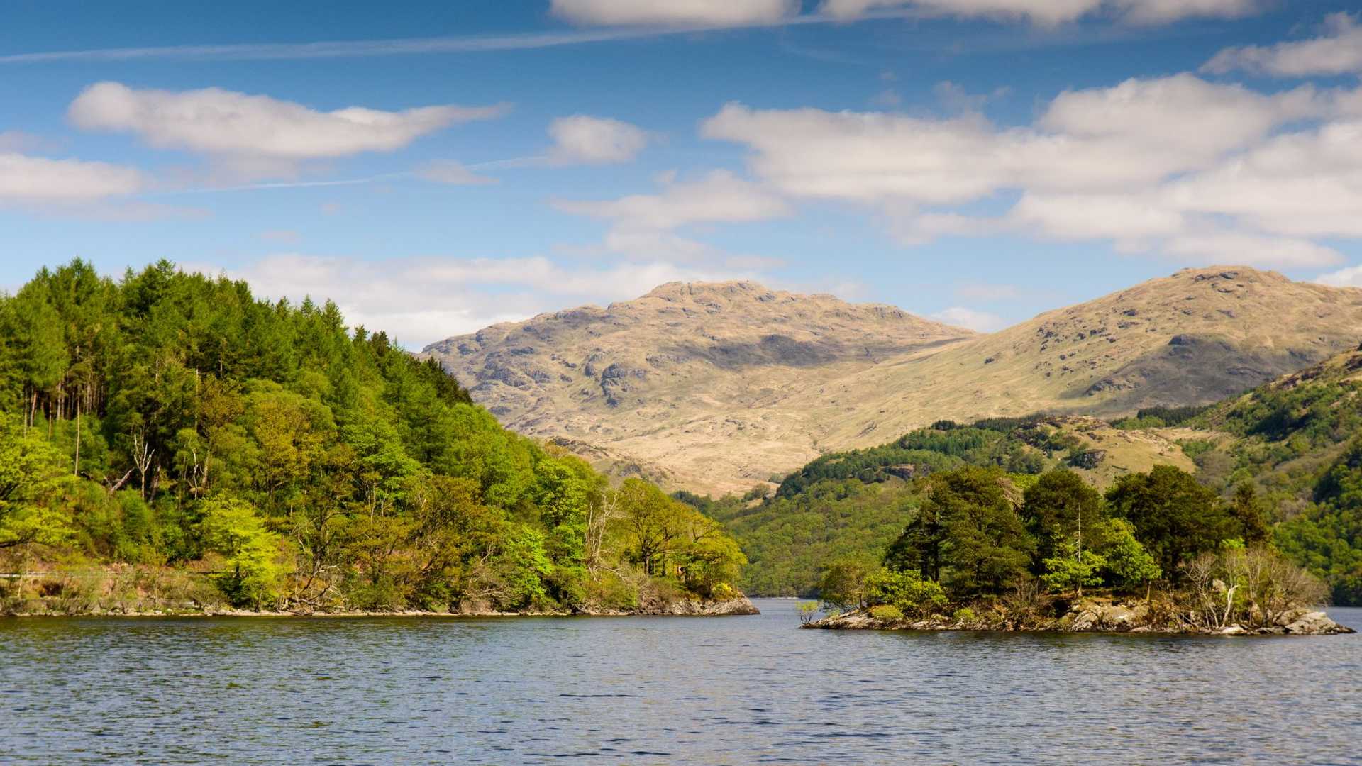 A wide body of water with a small, tree-covered island in the foreground, leading to lush green hills and barren, rocky mountains under a cloudy blue sky.