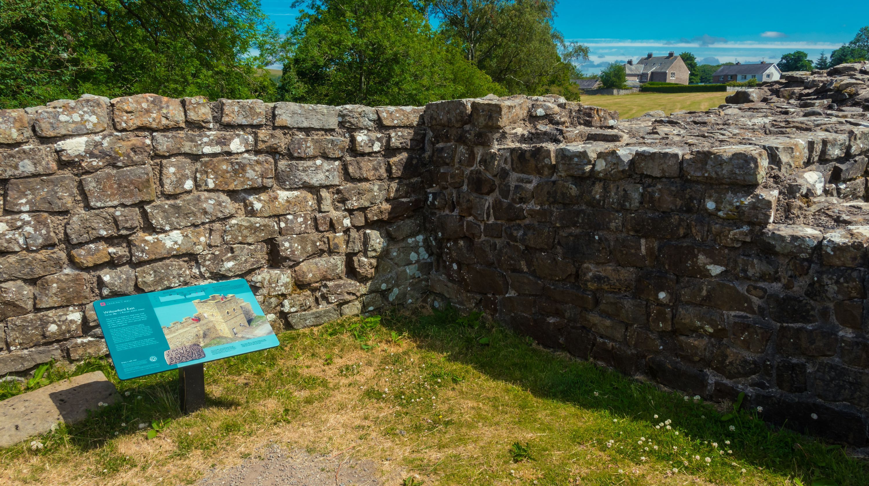 Stone ruins of a historical structure with an information sign in front of it, under a bright blue sky.
