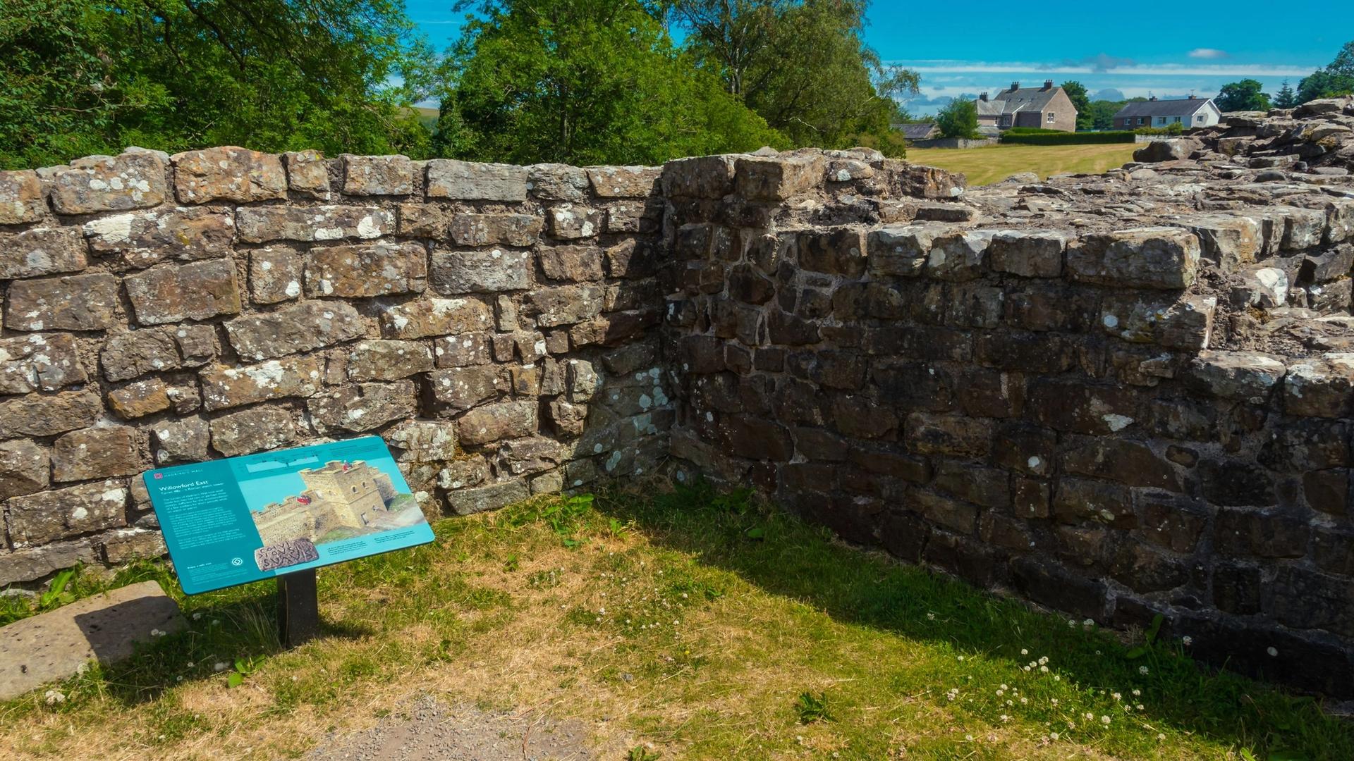 Stone ruins of a historical structure with an information sign in front of it, under a bright blue sky.