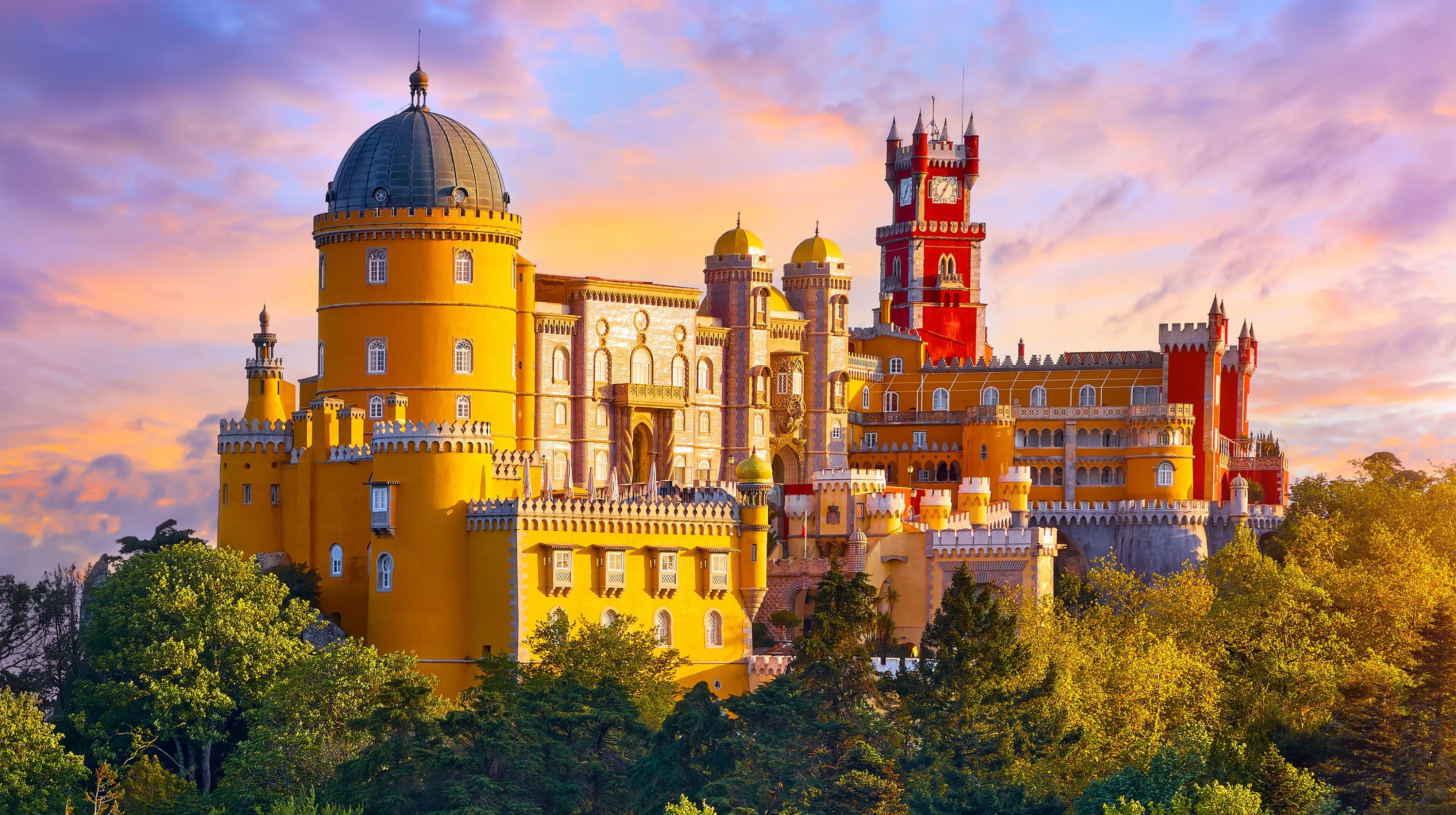 A vibrant yellow castle with a red tower, a domed turret, and intricate architecture, set against a colorful sunset sky and surrounded by lush green trees.