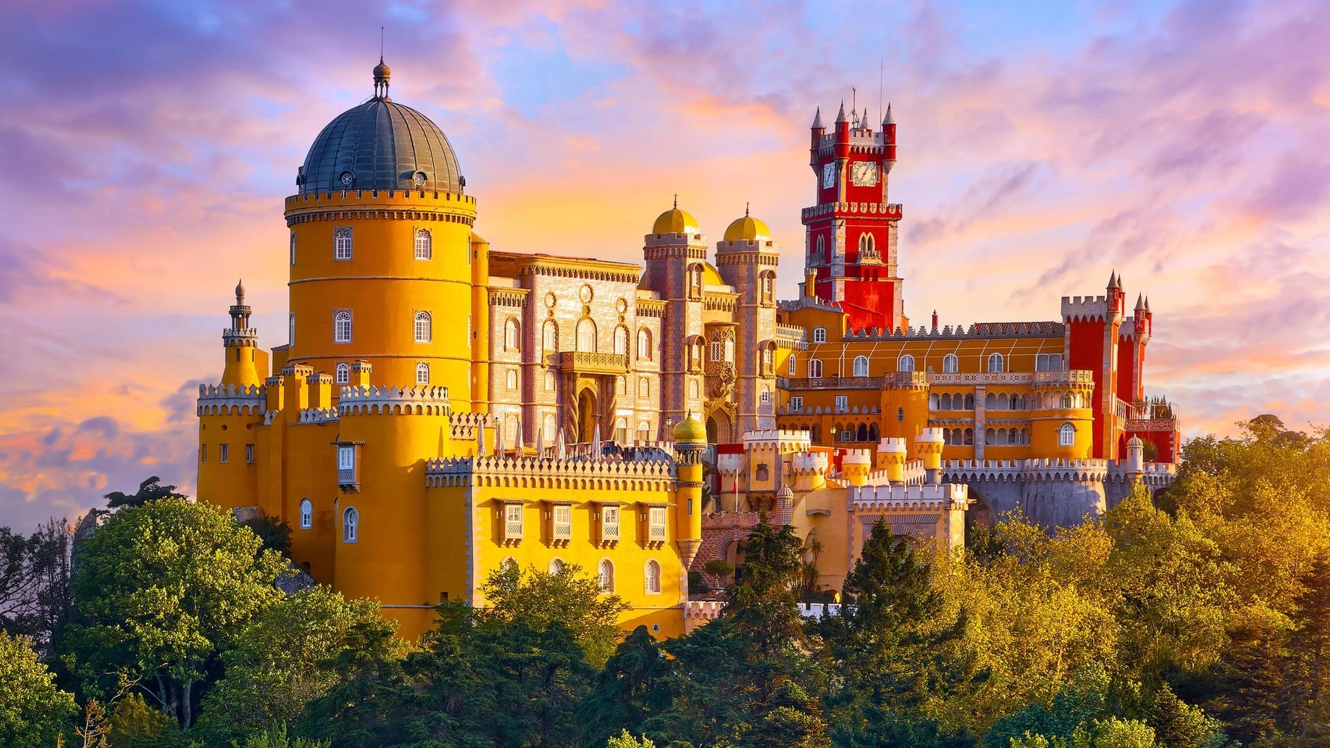 A vibrant yellow castle with a red tower, a domed turret, and intricate architecture, set against a colorful sunset sky and surrounded by lush green trees.