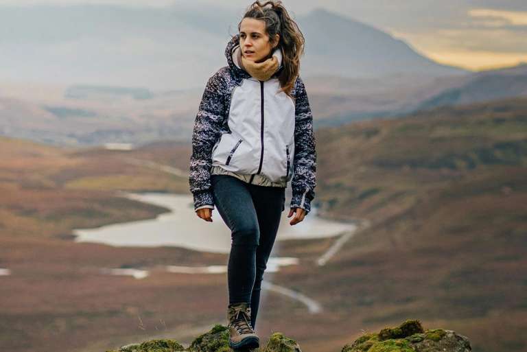 A person stands on a rocky outcrop overlooking a vast, mountainous landscape with a winding river below.