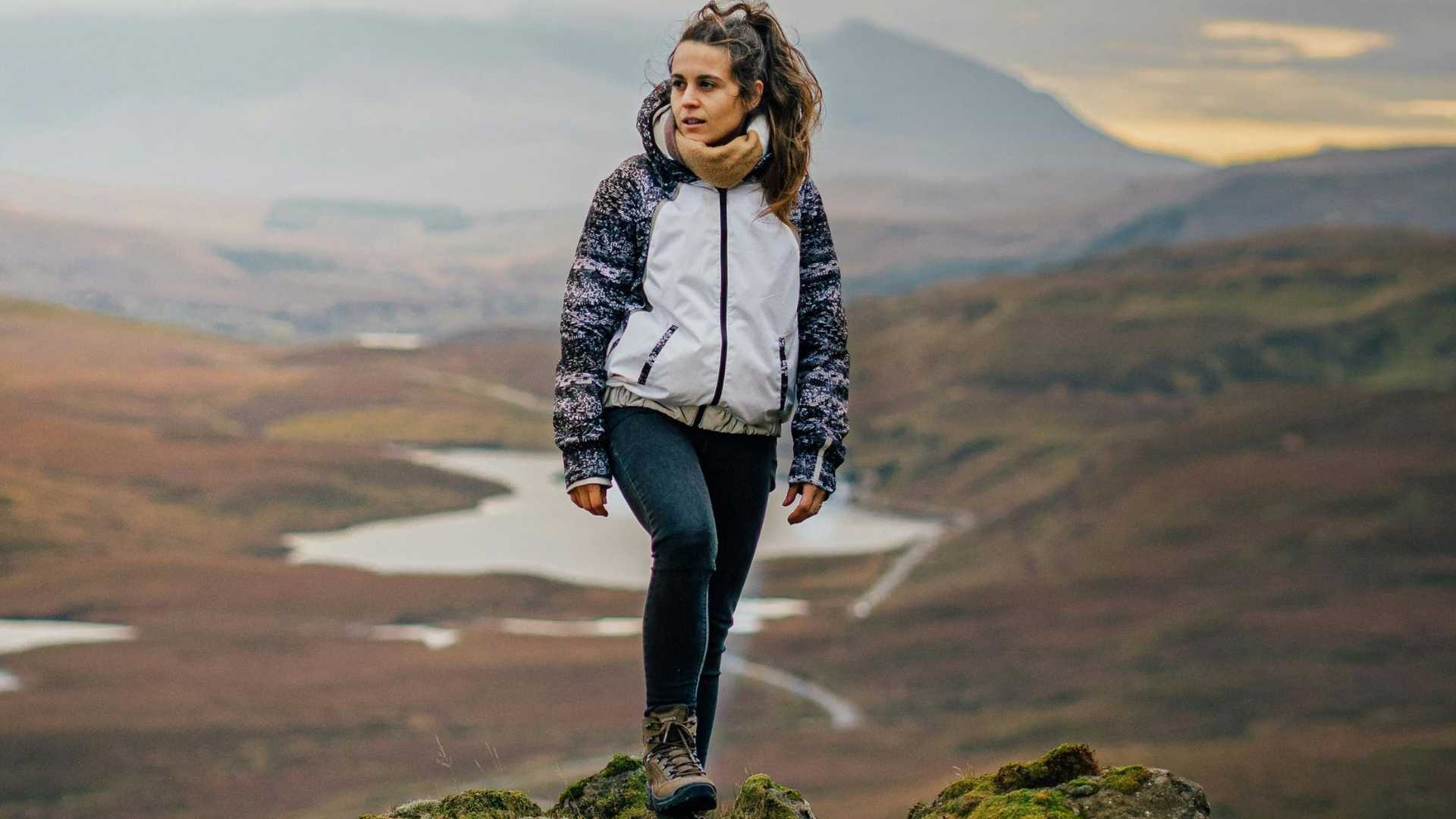A person stands on a rocky outcrop overlooking a vast, mountainous landscape with a winding river below.