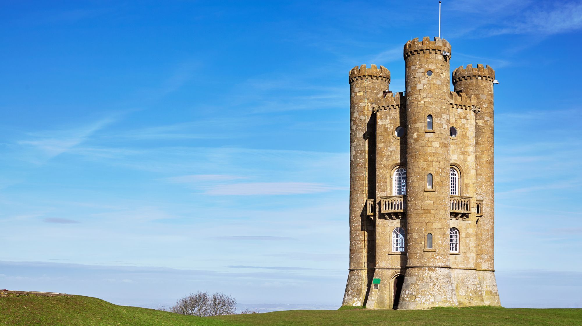 Broadway tower in the Cotswolds, with battlements and arched windows stands tall against a bright blue sky with wispy clouds.