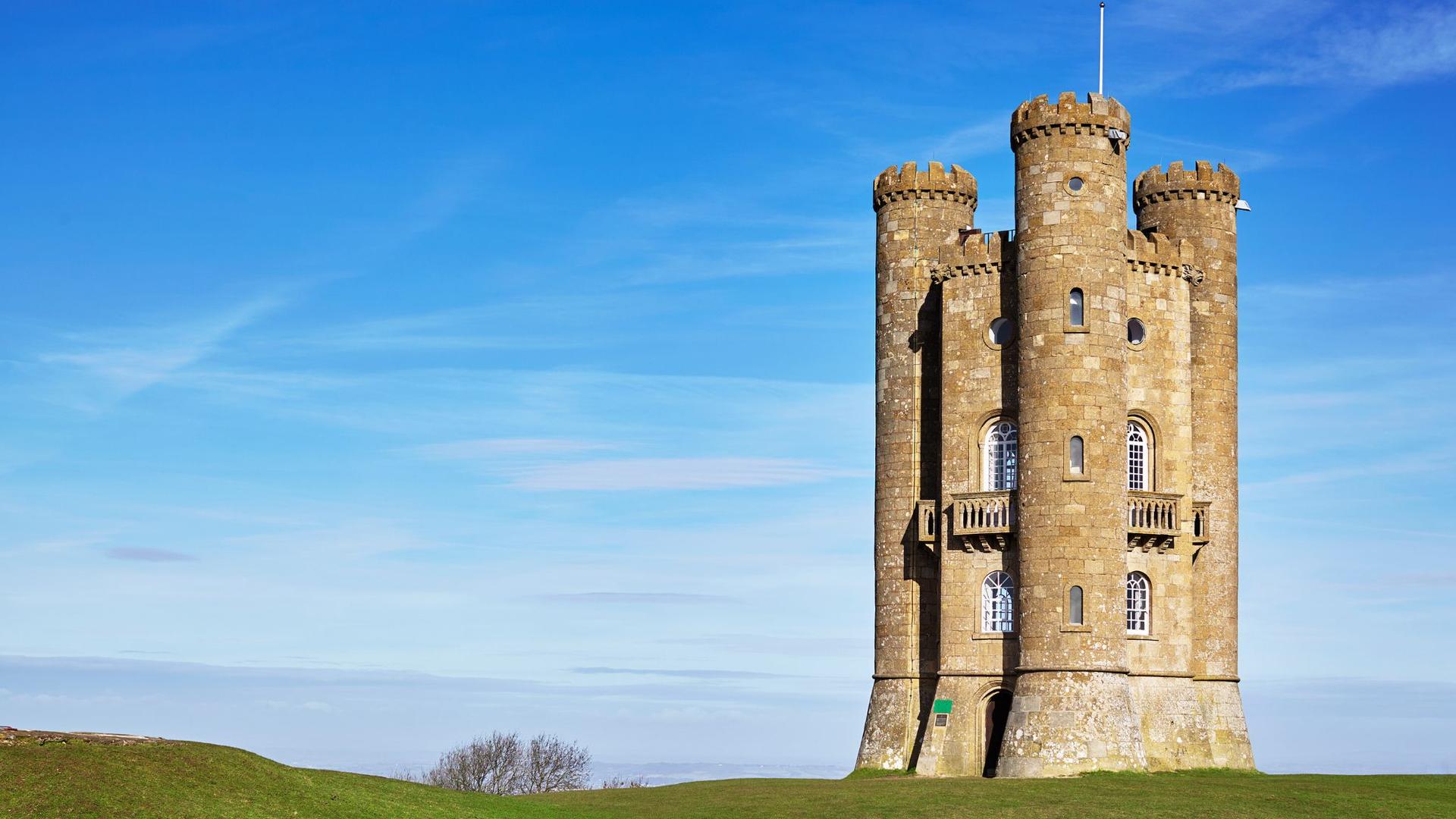 Broadway tower in the Cotswolds, with battlements and arched windows stands tall against a bright blue sky with wispy clouds.