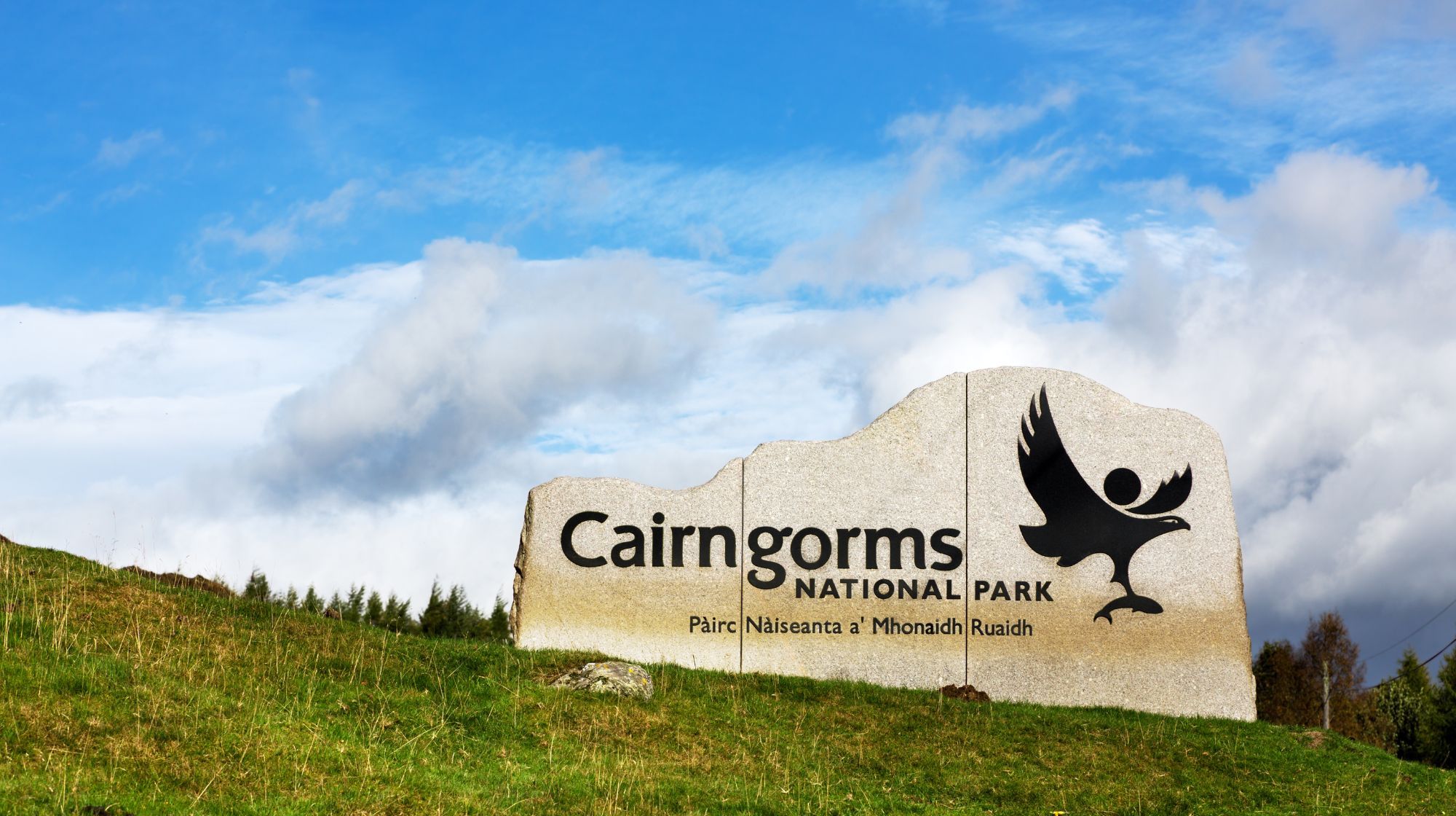 A stone monument at the Cairngorms National Park entrance features the park's name and a black silhouette of a bird in flight against a blue, cloudy sky.