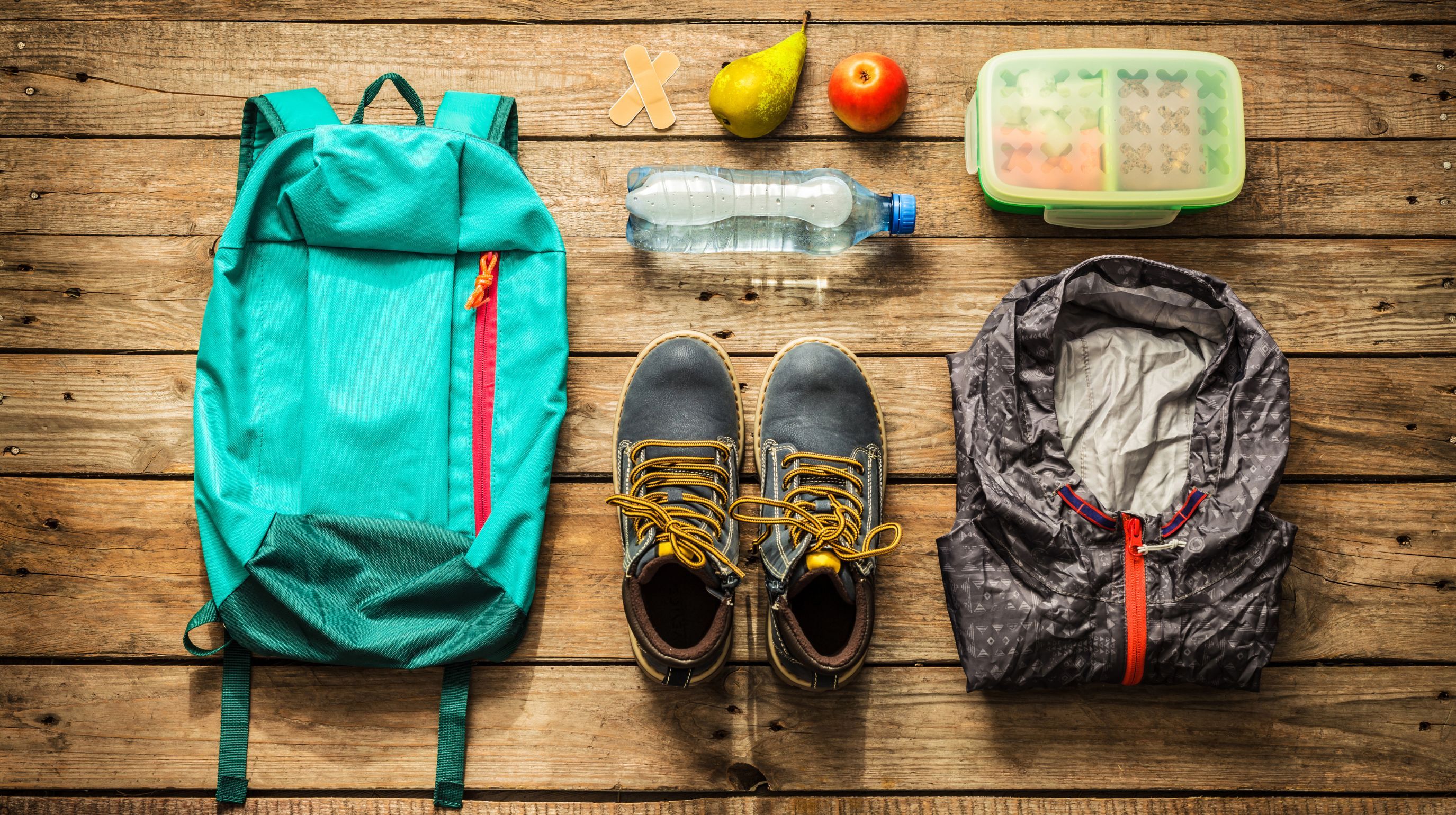 A teal backpack, a pair of boots, a jacket, a water bottle, a lunchbox, a pear, an apple, and two bandages are laid out on a wooden surface.