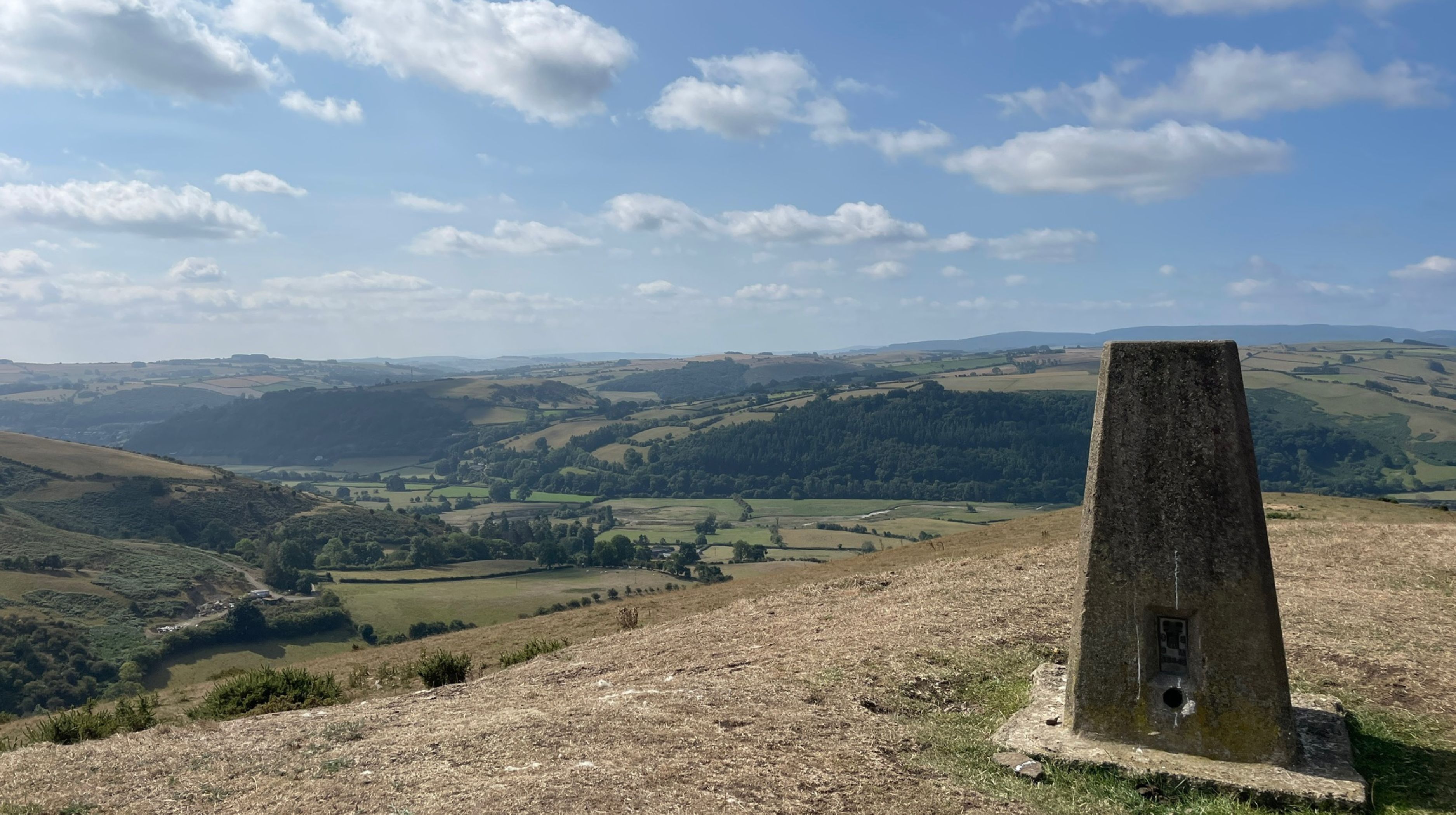 A concrete trig pillar stands on a dry, grassy hilltop overlooking a rolling valley with scattered trees and fields under a partly cloudy blue sky.