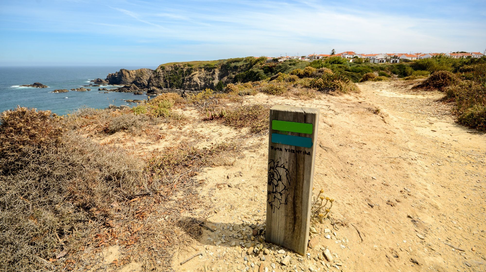 A wooden trail marker with green and blue stripes stands on a sandy path overlooking the ocean with rocky cliffs and a distant town.