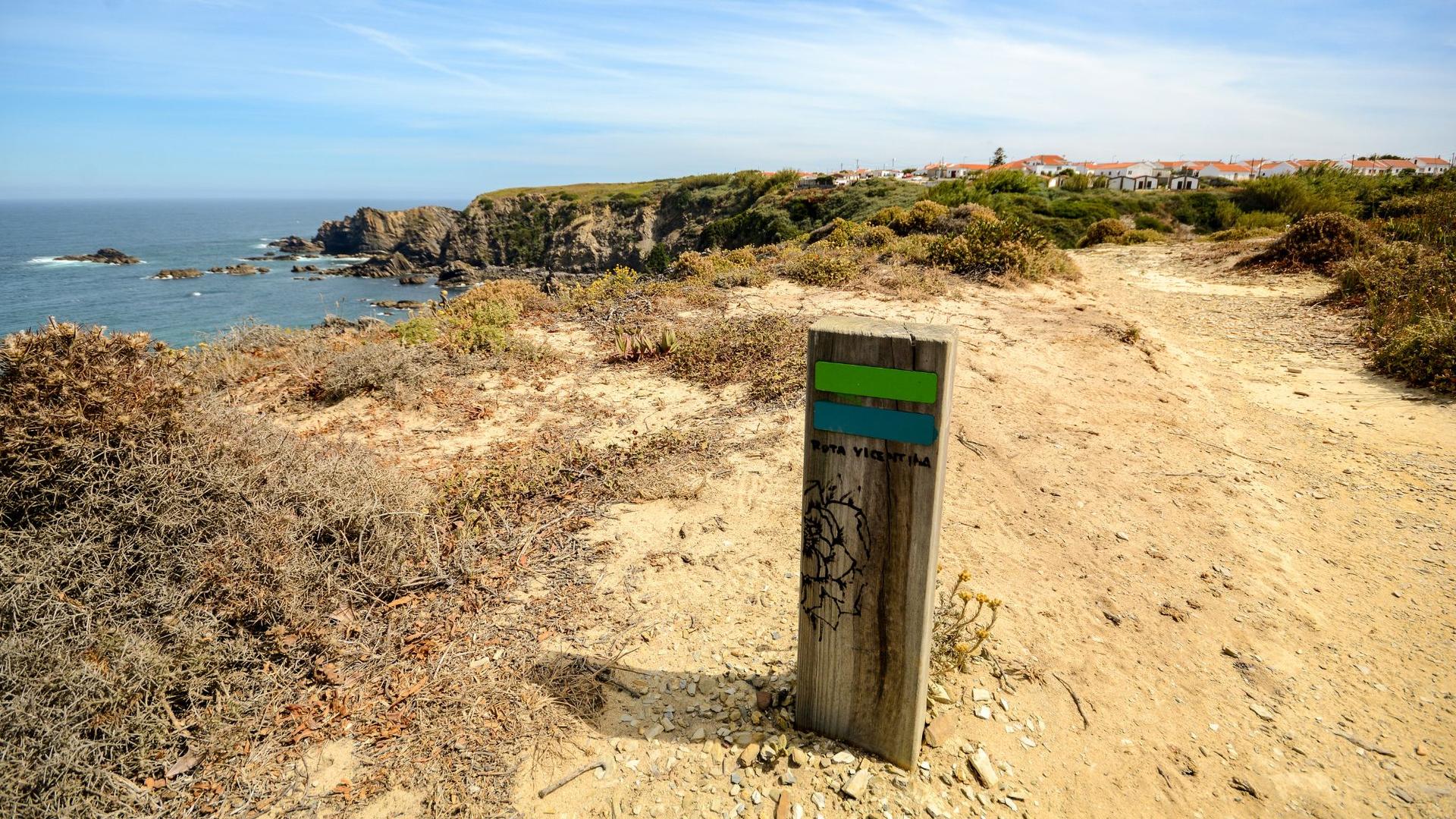 A wooden trail marker with green and blue stripes stands on a sandy path overlooking the ocean with rocky cliffs and a distant town.