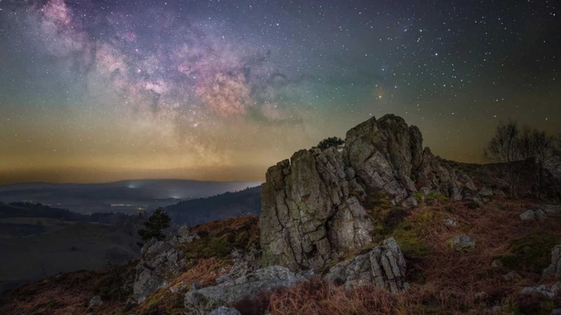 The Milky Way galaxy arches over a rocky, heather-covered hilltop with a valley and distant lights below.