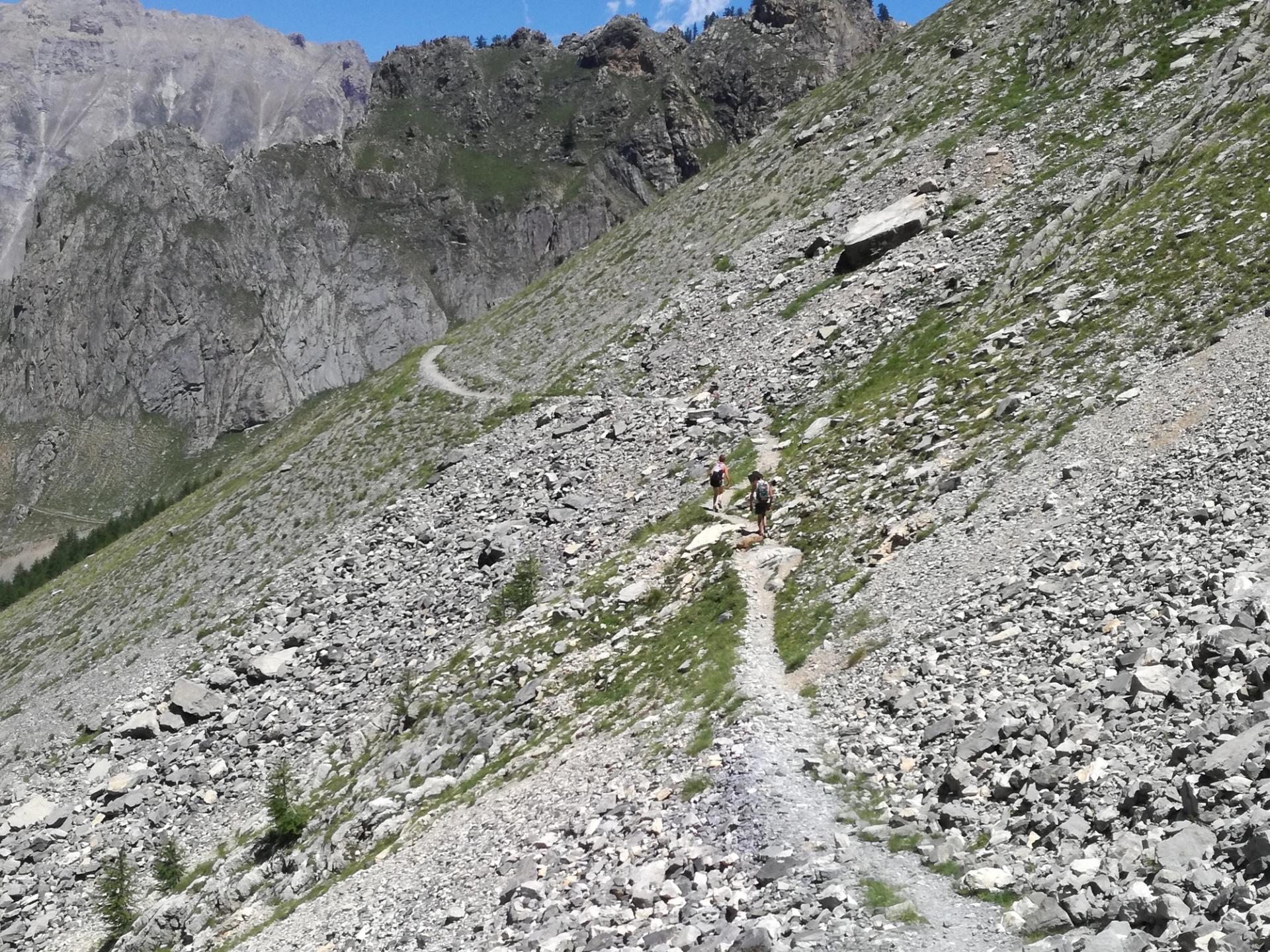 Two walkers following a narrow, stony mountain path beneath towering rock faces in Val Maira, Italy.