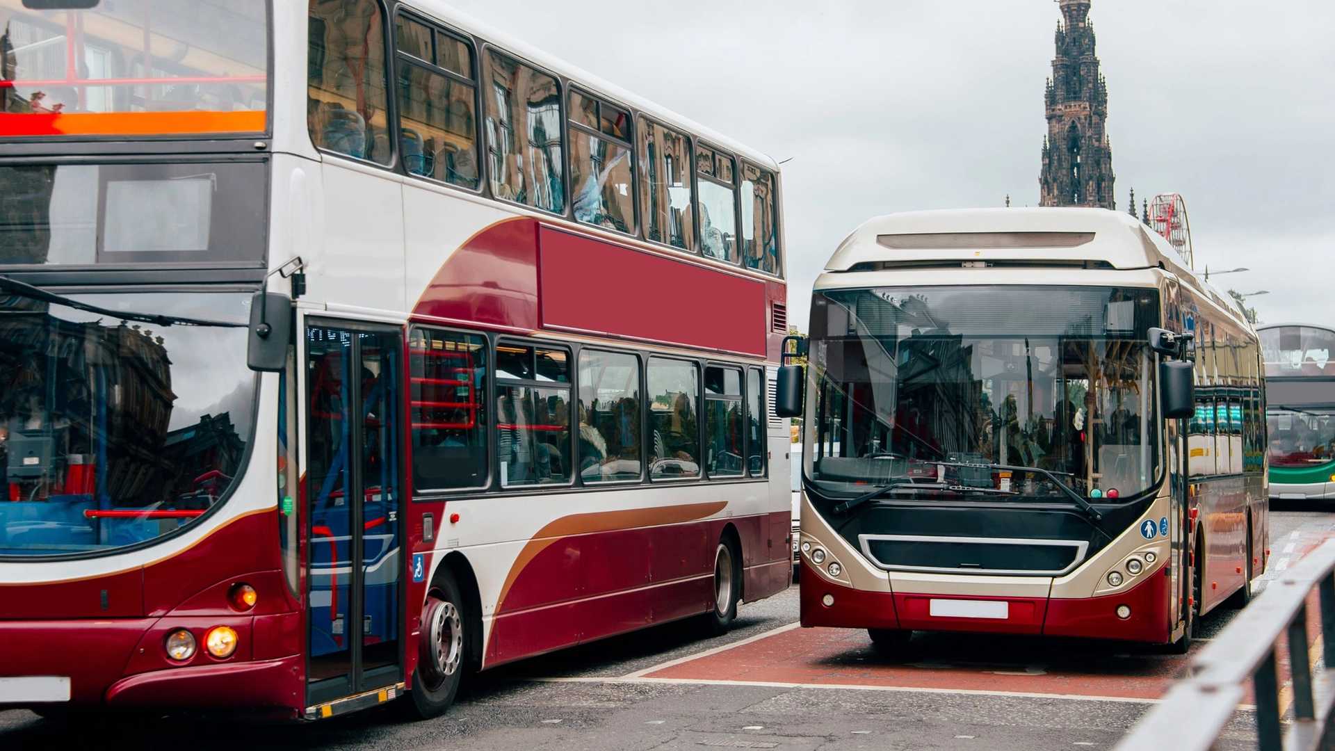 A red and white double-decker bus is parked next to a modern bus with a tall monument in the background.