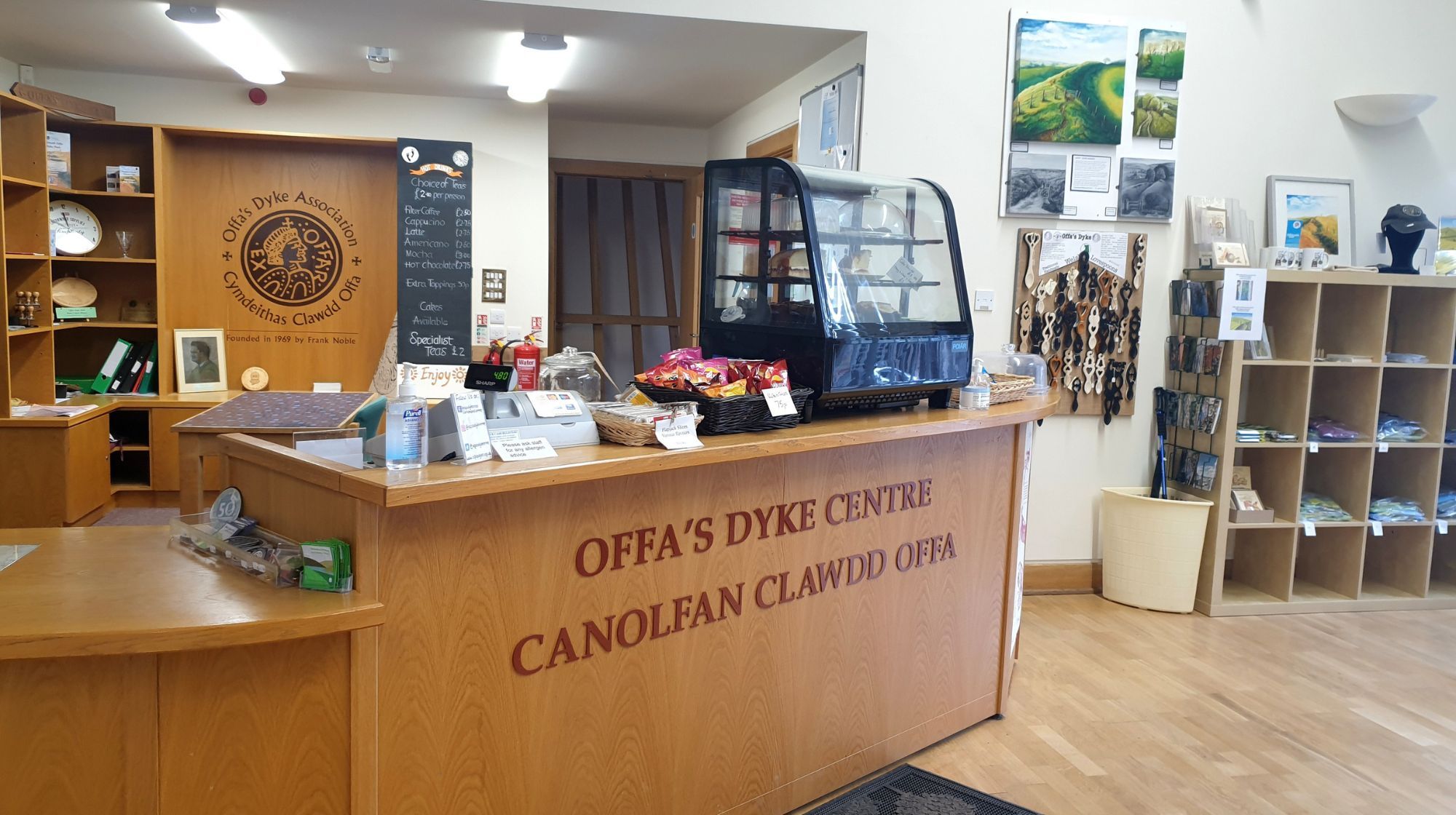 Interior of Offa’s Dyke Centre visitor reception, showing a wooden front desk with bilingual Welsh and English signage, a small café counter with snacks and a glass display case, and shelves of souvenirs and leaflets. The space is bright, tidy, and welcoming, with wooden floors and informational displays on the walls.