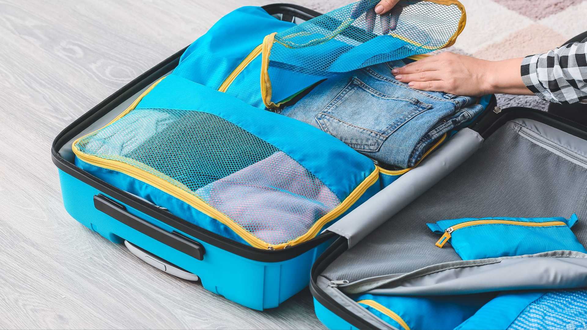 A person is packing a blue suitcase with clothing in mesh organizers. Jeans are visible inside one organiser.