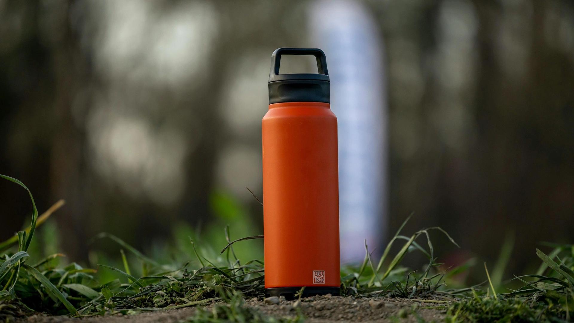 An orange insulated water bottle with a black lid and handle sits on the ground amidst green grass and blurred foliage.