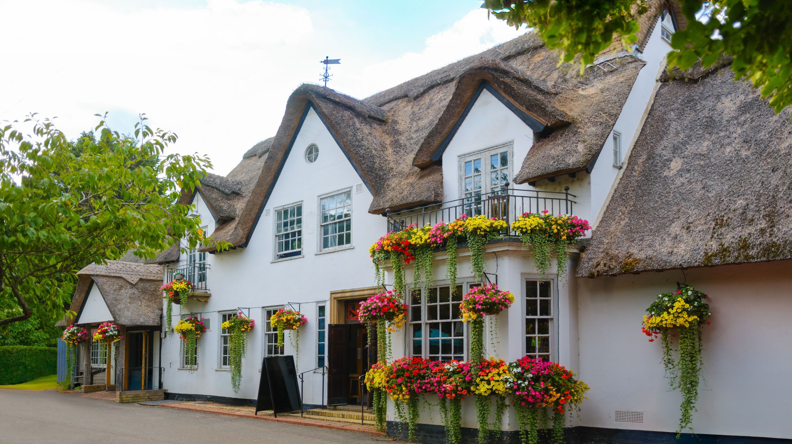 A white building with a thatched roof, adorned with numerous hanging baskets overflowing with colorful flowers.