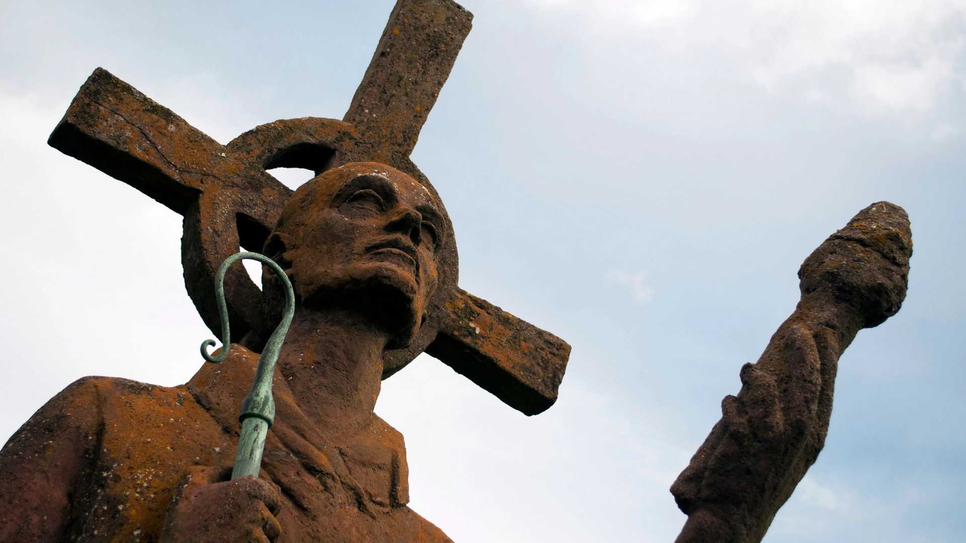 A stone statue of St Cuthbert holding a cross and a staff against a cloudy sky.