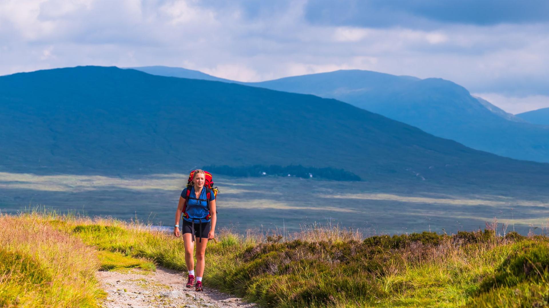 A person with a red backpack walks on a dirt path through grassy hills toward distant blue mountains under a cloudy sky.
