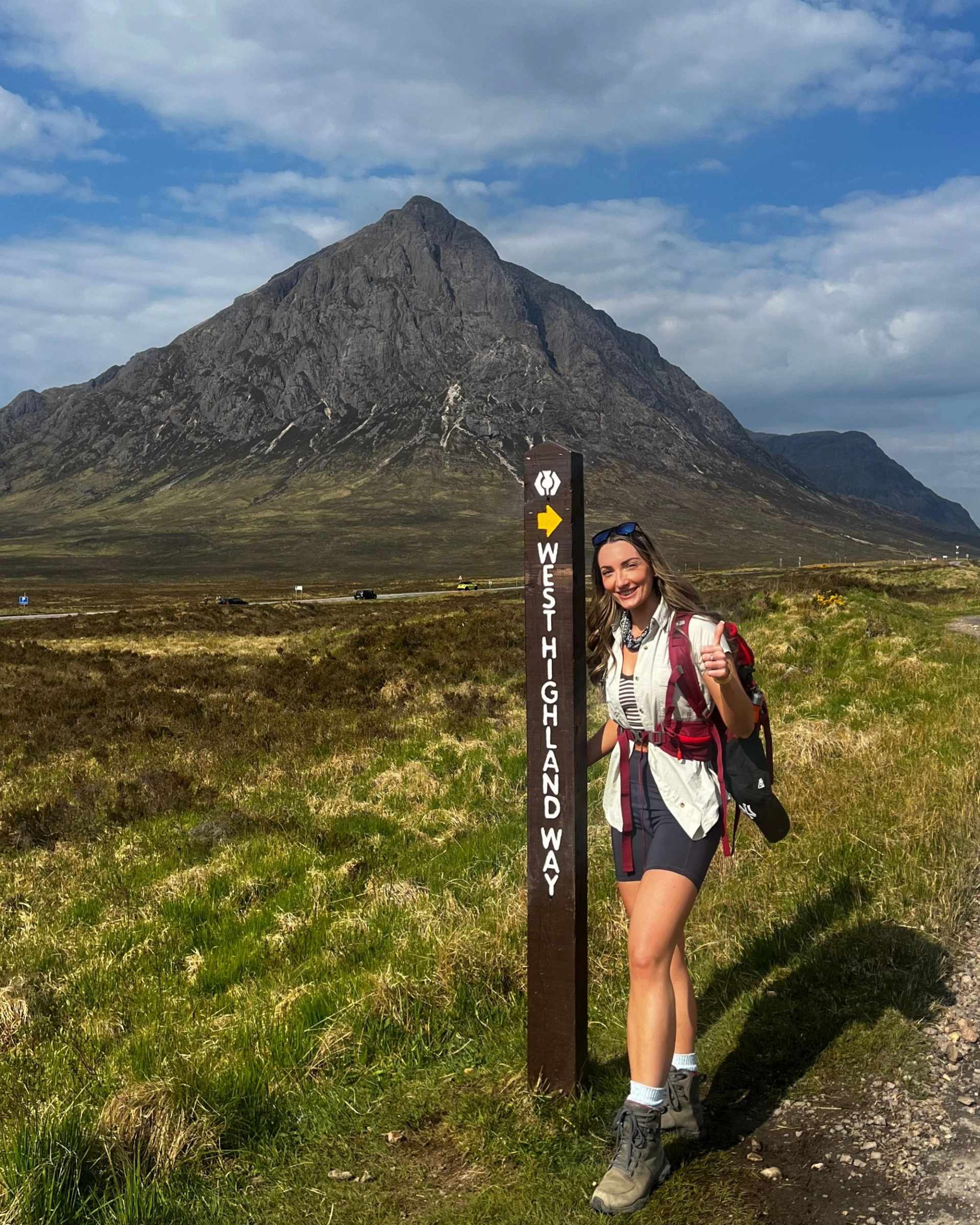 A hiker poses next to a West Highland Way signpost with a large mountain in the background